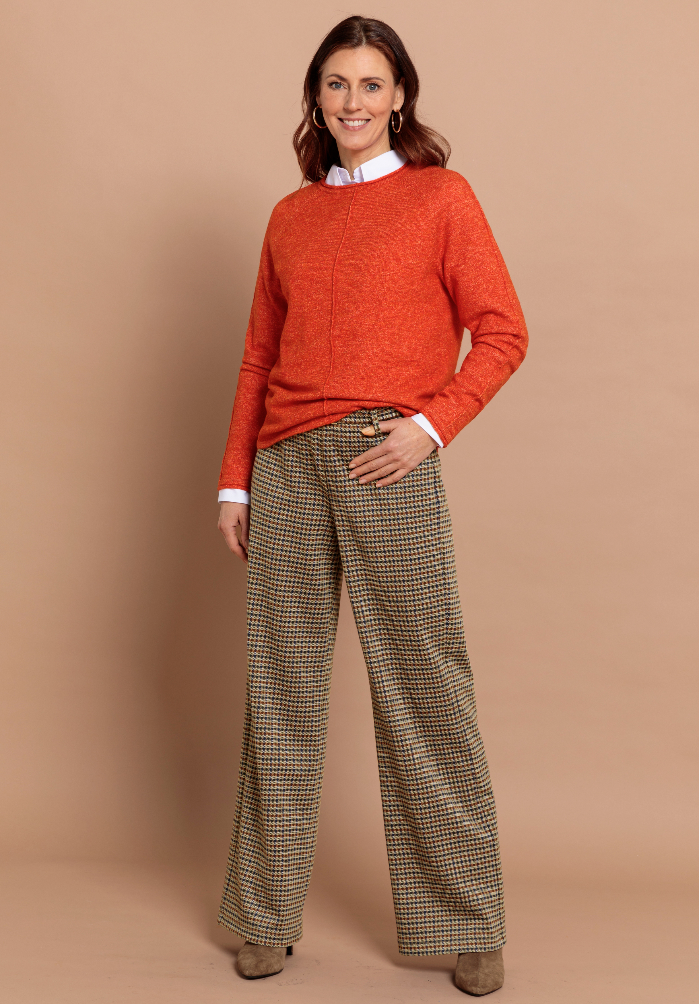 Eine Frau trägt einen leuchtend orangefarbenen Pullover über einem weißen Hemd mit Kragen, eine weite karierte Hose mit Hahnentrittmuster und braune Schuhe. Sie hat braunes Haar.