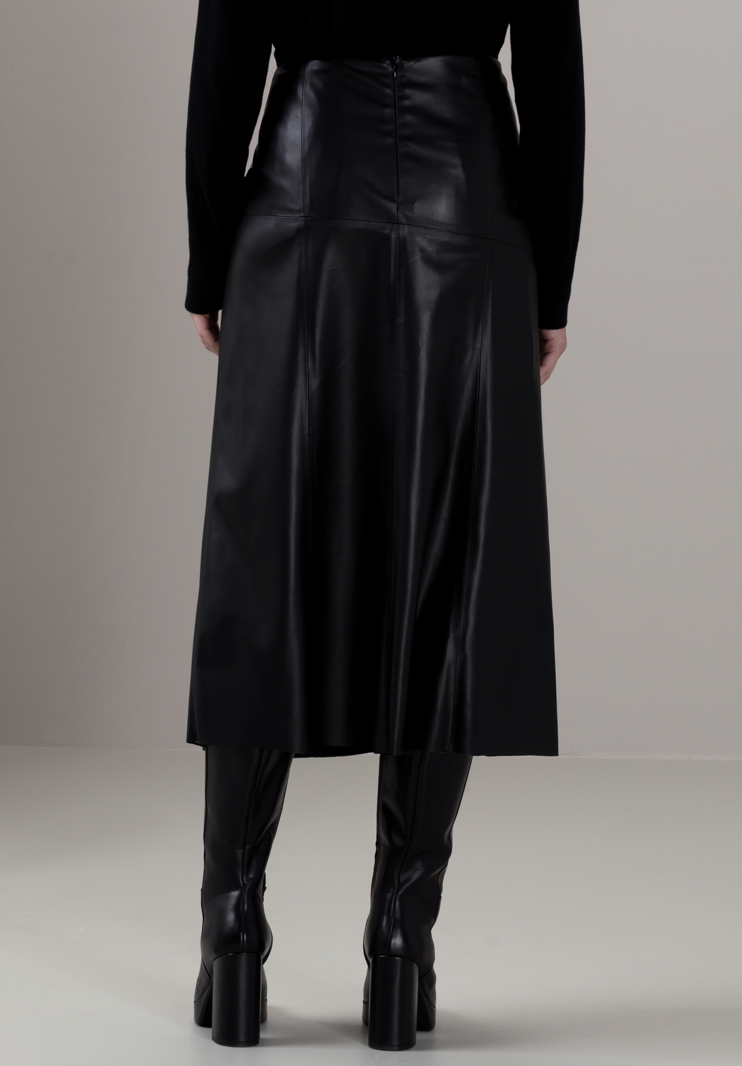 Eine Person, die ein schwarzes Langarmoberteil, einen hoch taillierten schwarzen Midirock aus Kunstleder und schwarze Stiefel mit Absatz trägt, von hinten vor einem schlichten hellen Hintergrund gezeigt.