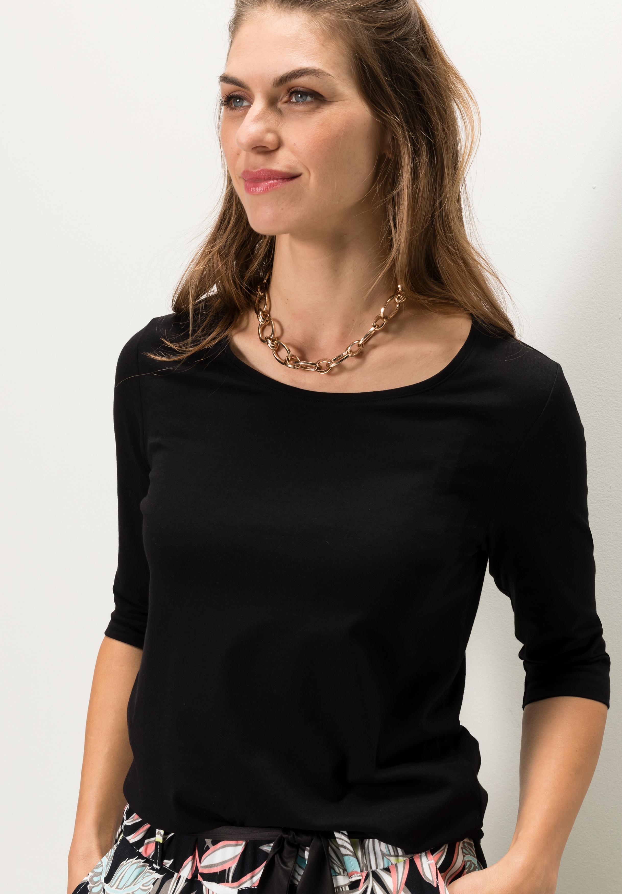 Eine Frau mit langen braunen Haaren, die ein schwarzes Oberteil mit dreiviertel Ärmeln, eine klobige Goldkette und eine gemusterte Hose trägt, steht vor einem schlichten weißen Hintergrund, blickt leicht zur Seite und lächelt sanft.