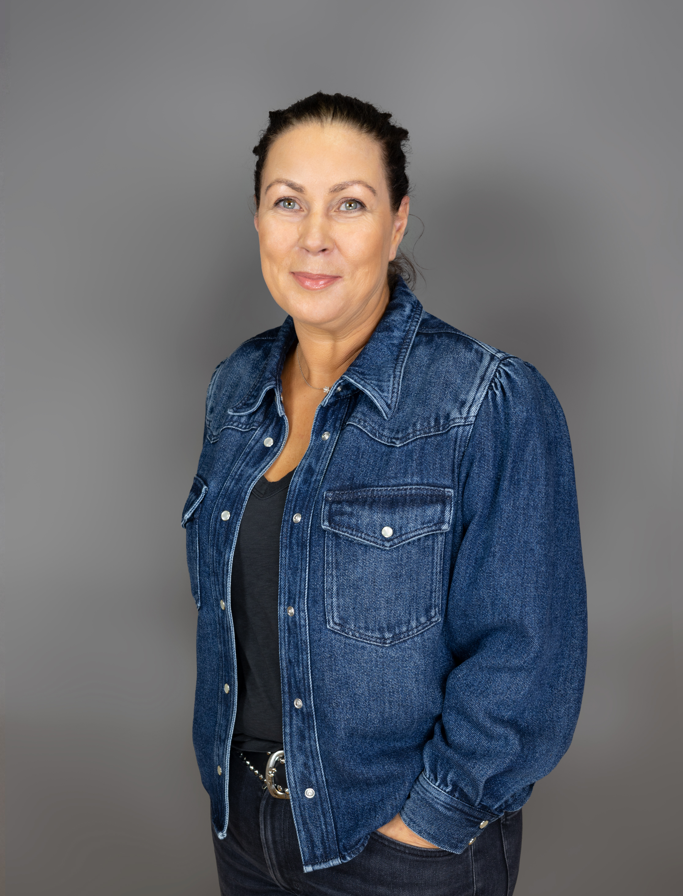 Eine Frau mit zurückgebundenem dunklem Haar und einer blauen Jeansjacke über einem schwarzen Oberteil steht vor einem schlichten grauen Hintergrund und lächelt sanft, die Hand in der Tasche.