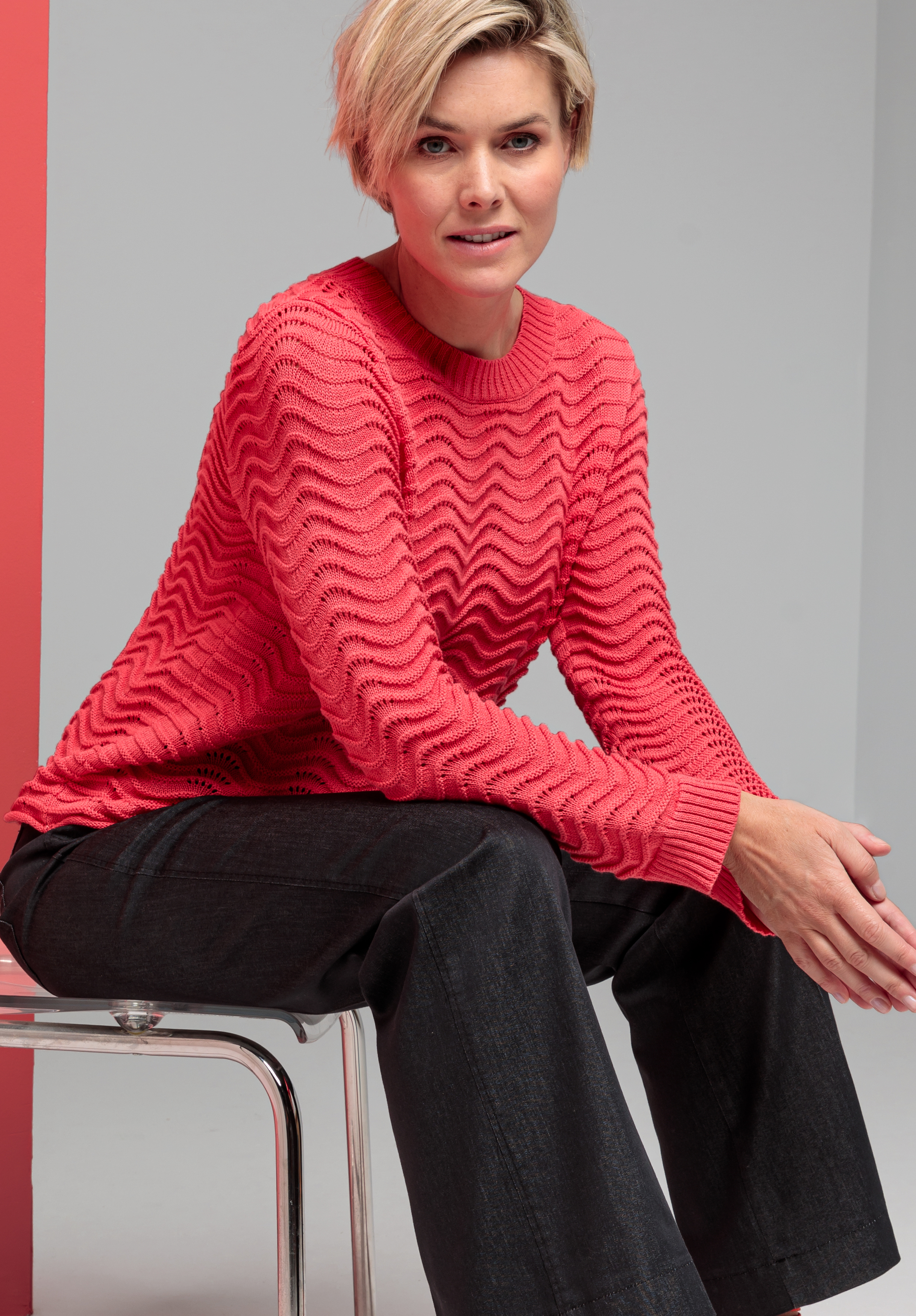 Eine Frau mit kurzen blonden Haaren sitzt auf einem durchsichtigen Stuhl. Sie trägt einen strukturierten, korallenfarbenen Pullover und eine schwarze Hose vor einem grau-roten Hintergrund.