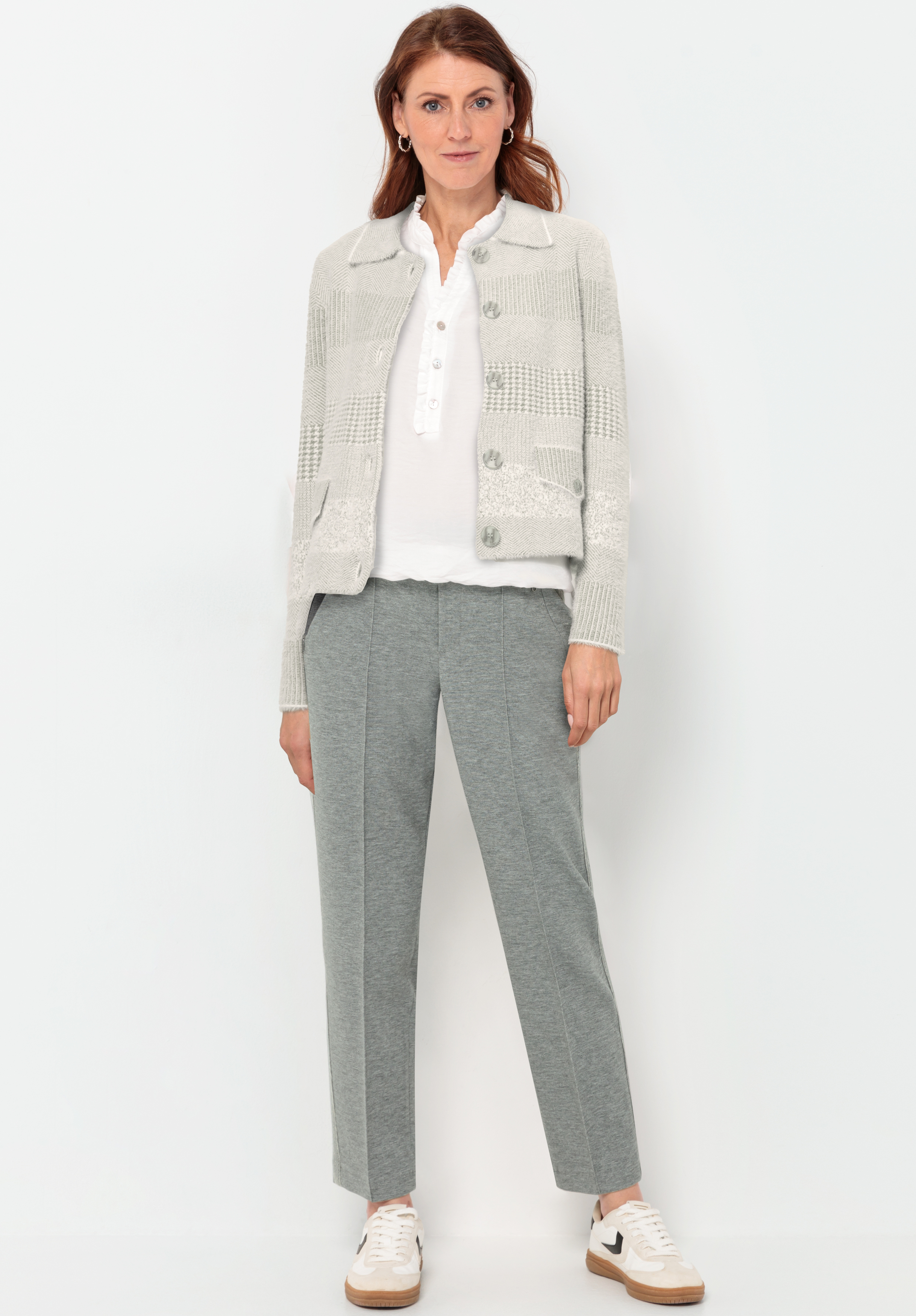 Eine Frau steht vor einem weißen Hintergrund und trägt eine hellgraue strukturierte Strickjacke, eine weiße Bluse mit Kragen, eine graue maßgeschneiderte Hose und weiße Turnschuhe mit beigen Akzenten. Sie schaut in die Kamera und lächelt leicht.