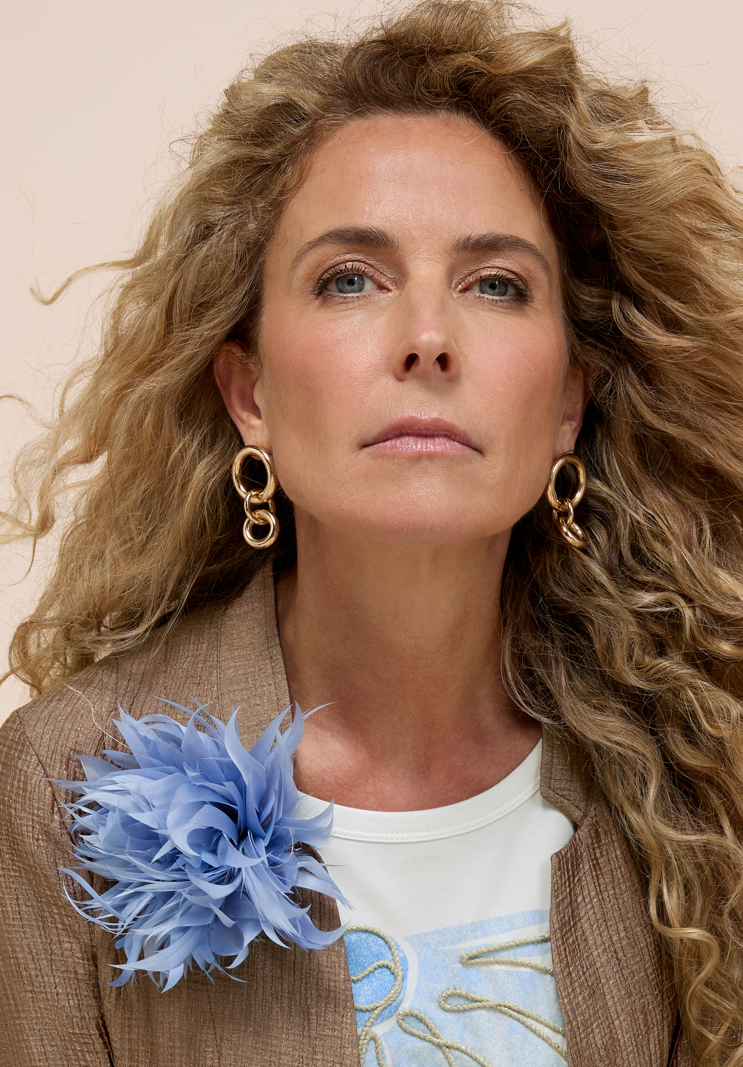 Eine Frau mit langen, lockigen blonden Haaren trägt goldene Creolen, eine braune, strukturierte Jacke und ein weißes Hemd mit einem blauen Aufdruck. An ihrer Jacke ist eine große, hellblaue Blumenbrosche befestigt. Sie blickt selbstbewusst in die Kamera.