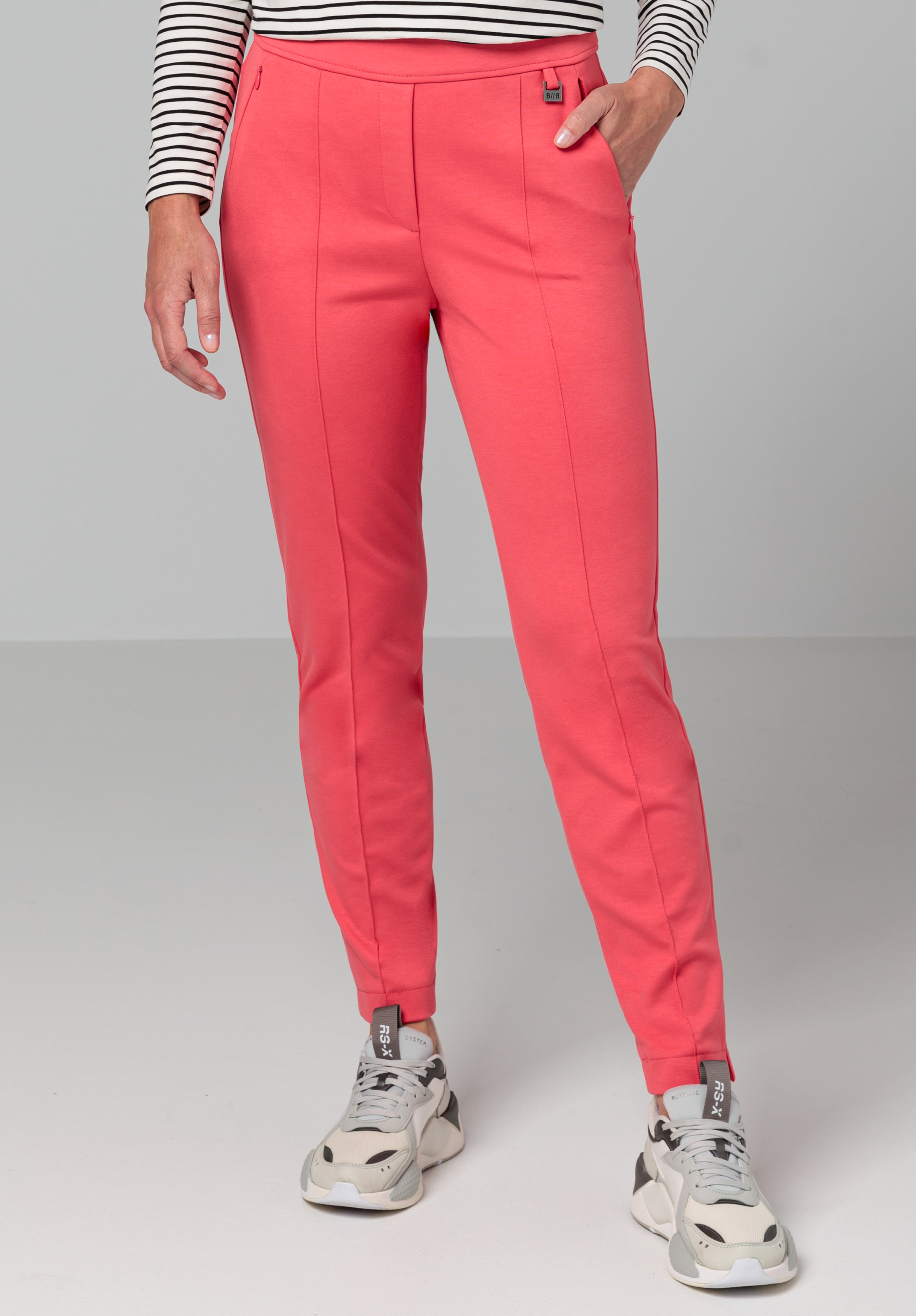 Eine Person trägt korallenrosa Hosen, ein schwarz-weiß gestreiftes Langarmoberteil und weiß-graue Turnschuhe und steht mit einer Hand in der Tasche vor einem schlichten Hintergrund.