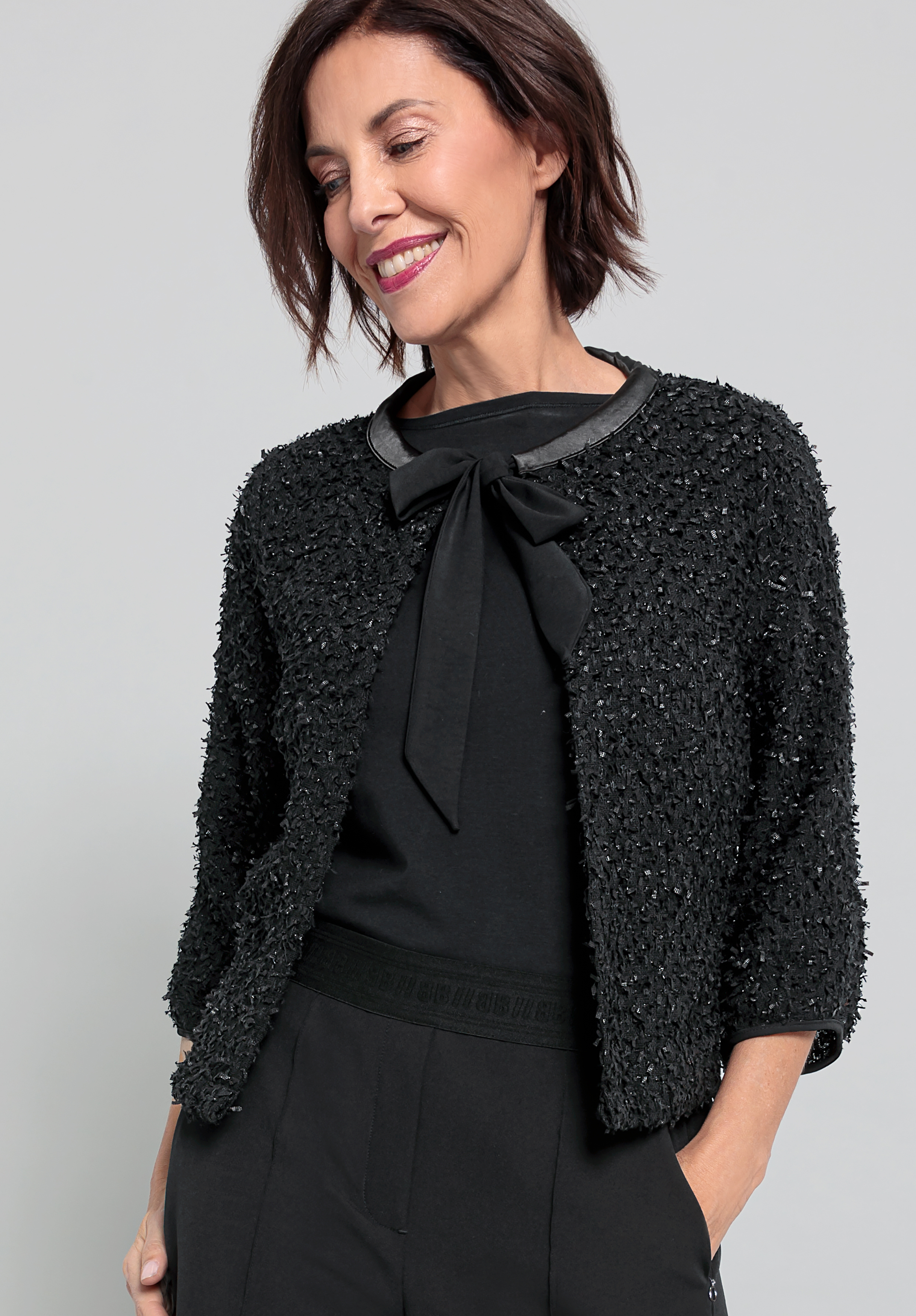 Eine Frau mit kurzen braunen Haaren lächelt, während sie eine strukturierte schwarze Jacke über einem schwarzen Oberteil mit einer Schleife am Ausschnitt und einer schwarzen Hose trägt; sie posiert mit den Händen in den Taschen vor einem hellgrauen Hinter