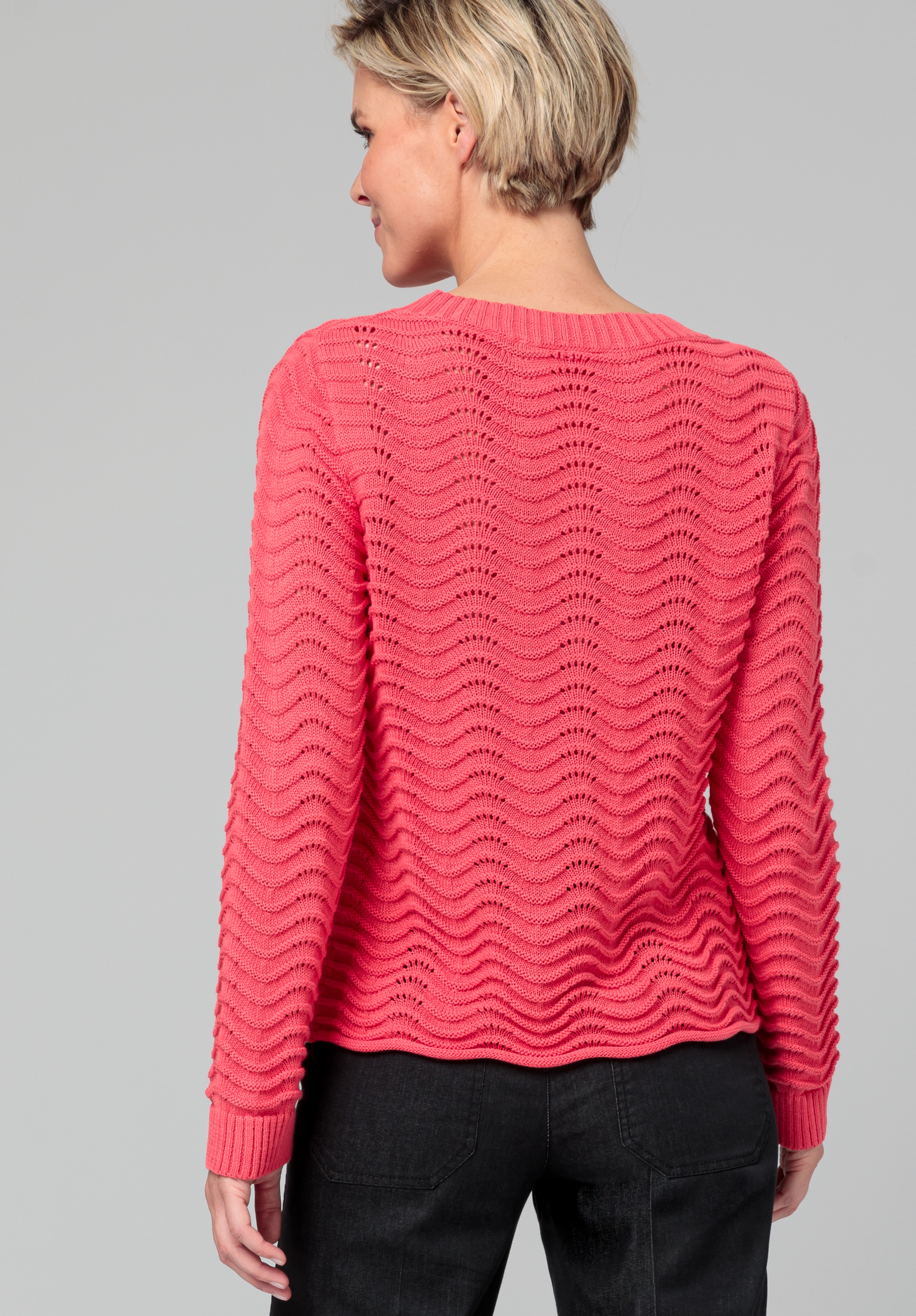 Von hinten ist eine Person mit kurzen blonden Haaren zu sehen, die einen strukturierten, langärmeligen, rosa Pullover mit gewelltem Strickmuster und eine schwarze Hose vor einem schlichten grauen Hintergrund trägt.