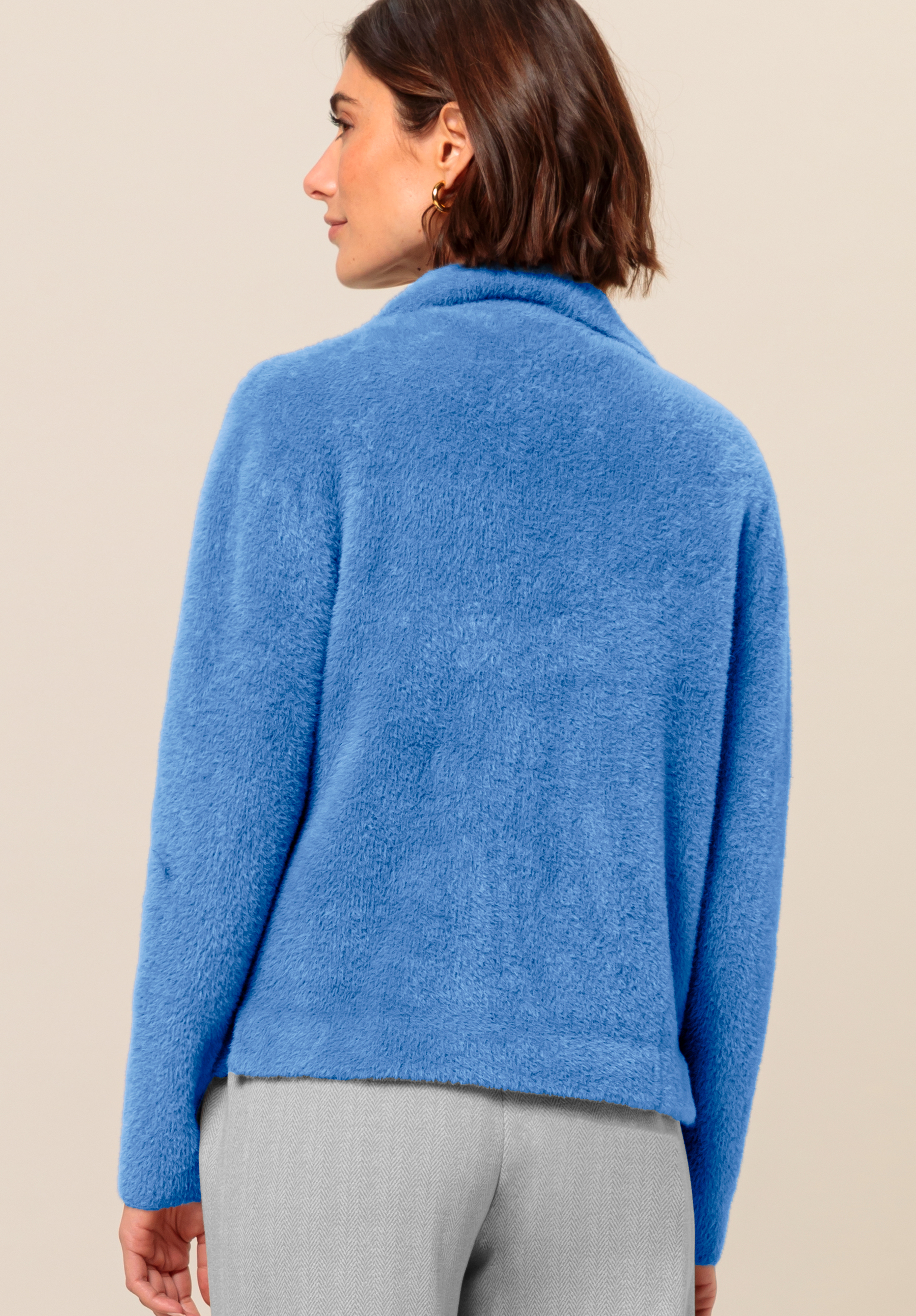 Eine Frau mit kurzen braunen Haaren, von hinten gesehen, trägt eine weiche, flauschige blaue Jacke und hellgraue Hosen vor einem schlichten beigen Hintergrund.