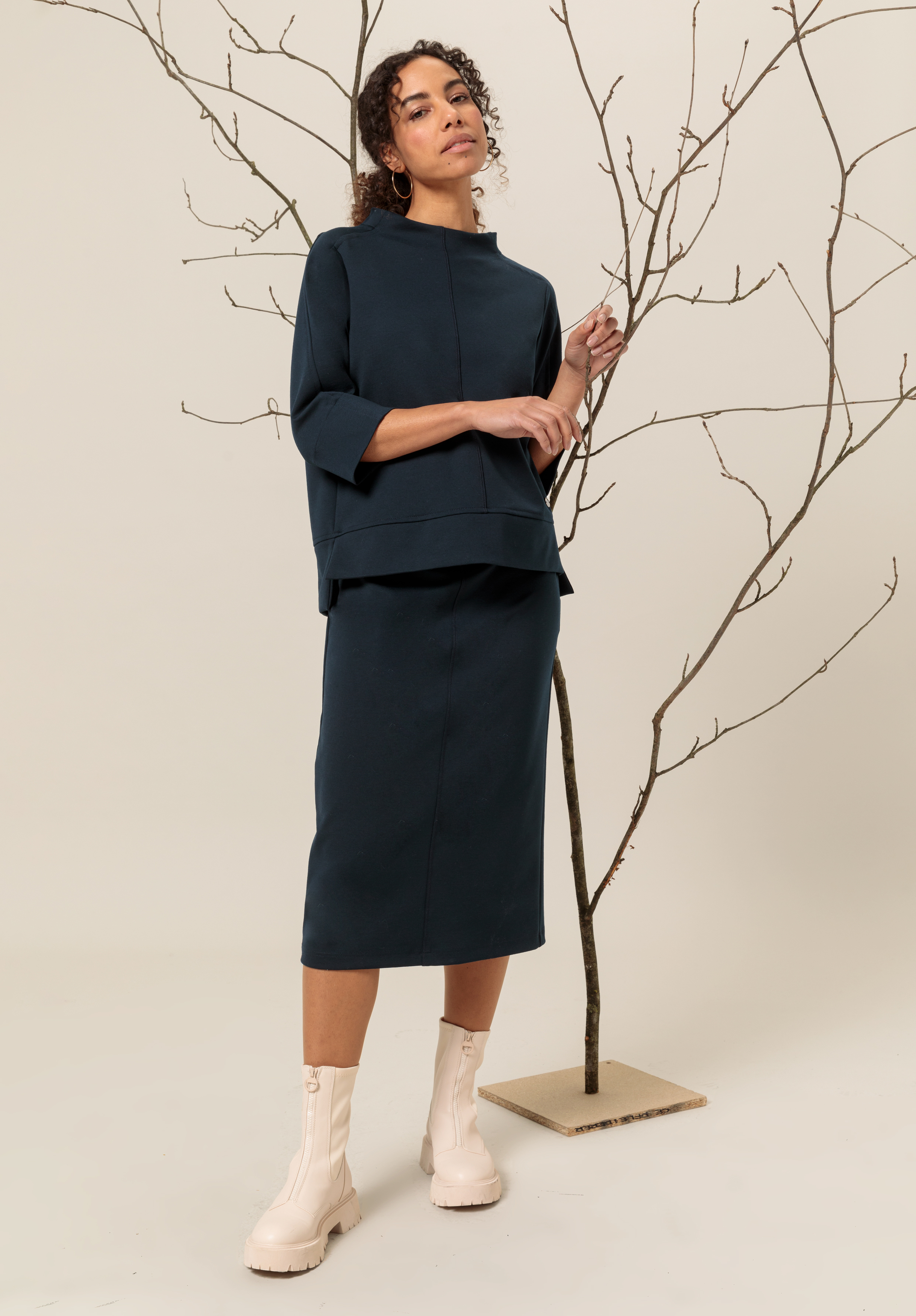 Eine Frau steht drinnen und trägt ein dunkelblaues Langarmshirt, einen passenden Midirock und hellbeige Stiefeletten. Sie posiert neben einem kahlen Ast vor einem neutralen Hintergrund.
