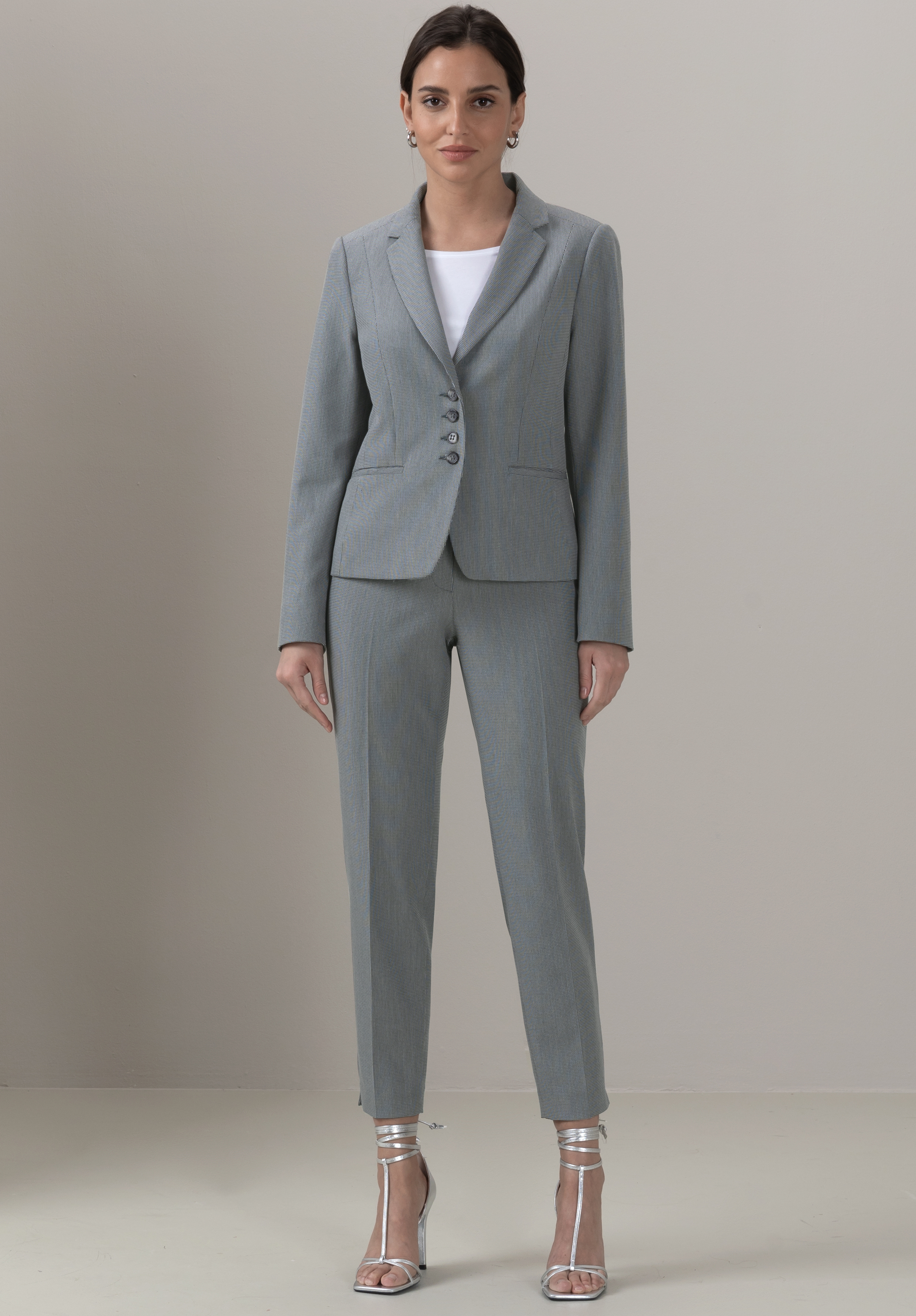Eine Frau steht mit dem Gesicht nach vorne, trägt einen hellgrauen maßgeschneiderten Hosenanzug mit einem weißen Oberteil darunter und silberne hochhackige Sandalen. Sie steht vor einem schlichten hellgrauen Hintergrund.