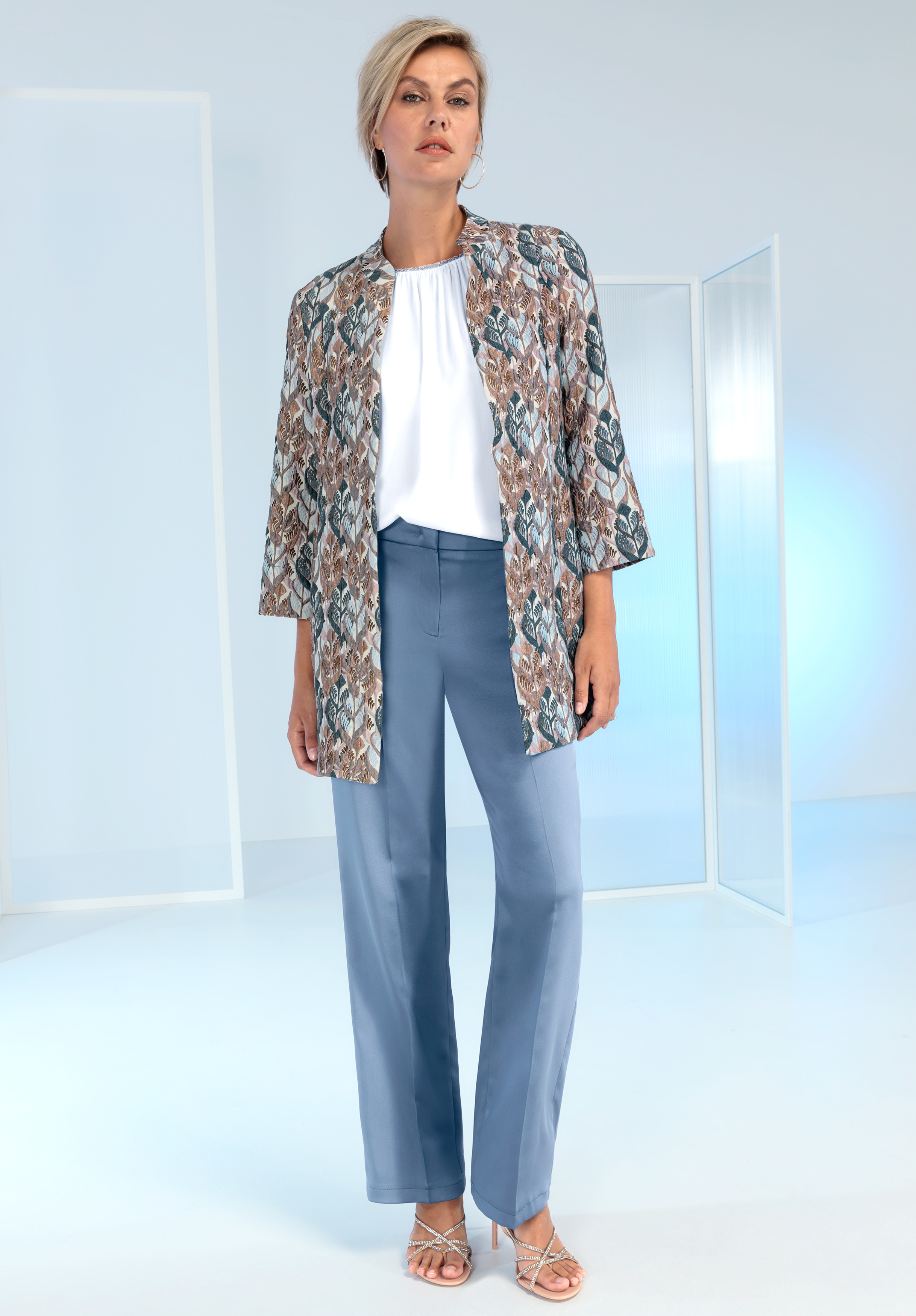 Eine Frau steht in einem minimalistisch-modernen Ambiente. Sie trägt eine hellblaue Hose, eine weiße Bluse, eine gemusterte offene Jacke in Erdtönen und Riemchensandalen. Sie hat kurze blonde Haare und einen selbstbewussten Gesichtsausdruck.