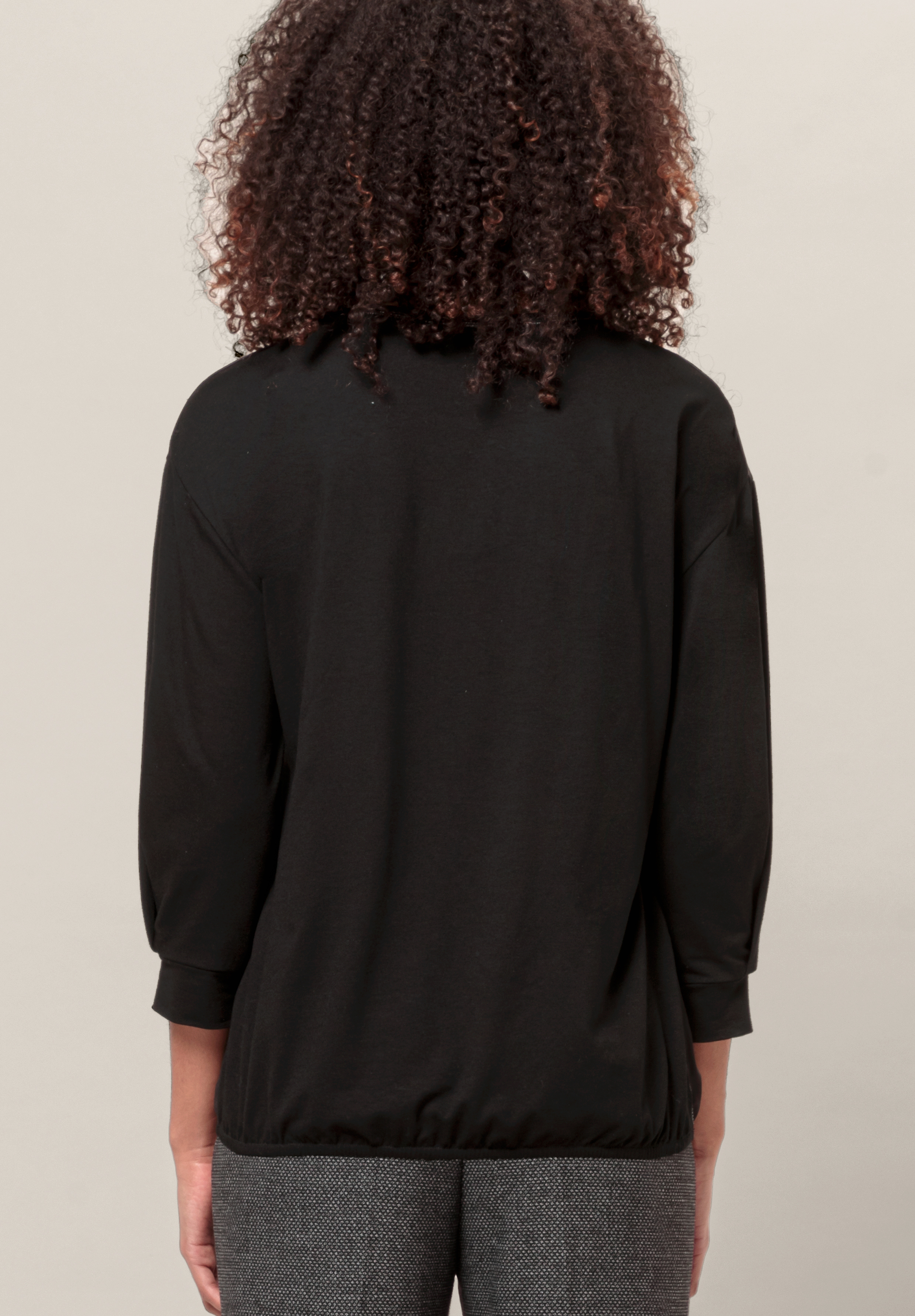 Von hinten ist eine Person mit lockigem Haar zu sehen, die vor einem schlichten hellen Hintergrund ein locker sitzendes schwarzes Langarmshirt und eine graue Hose trägt.