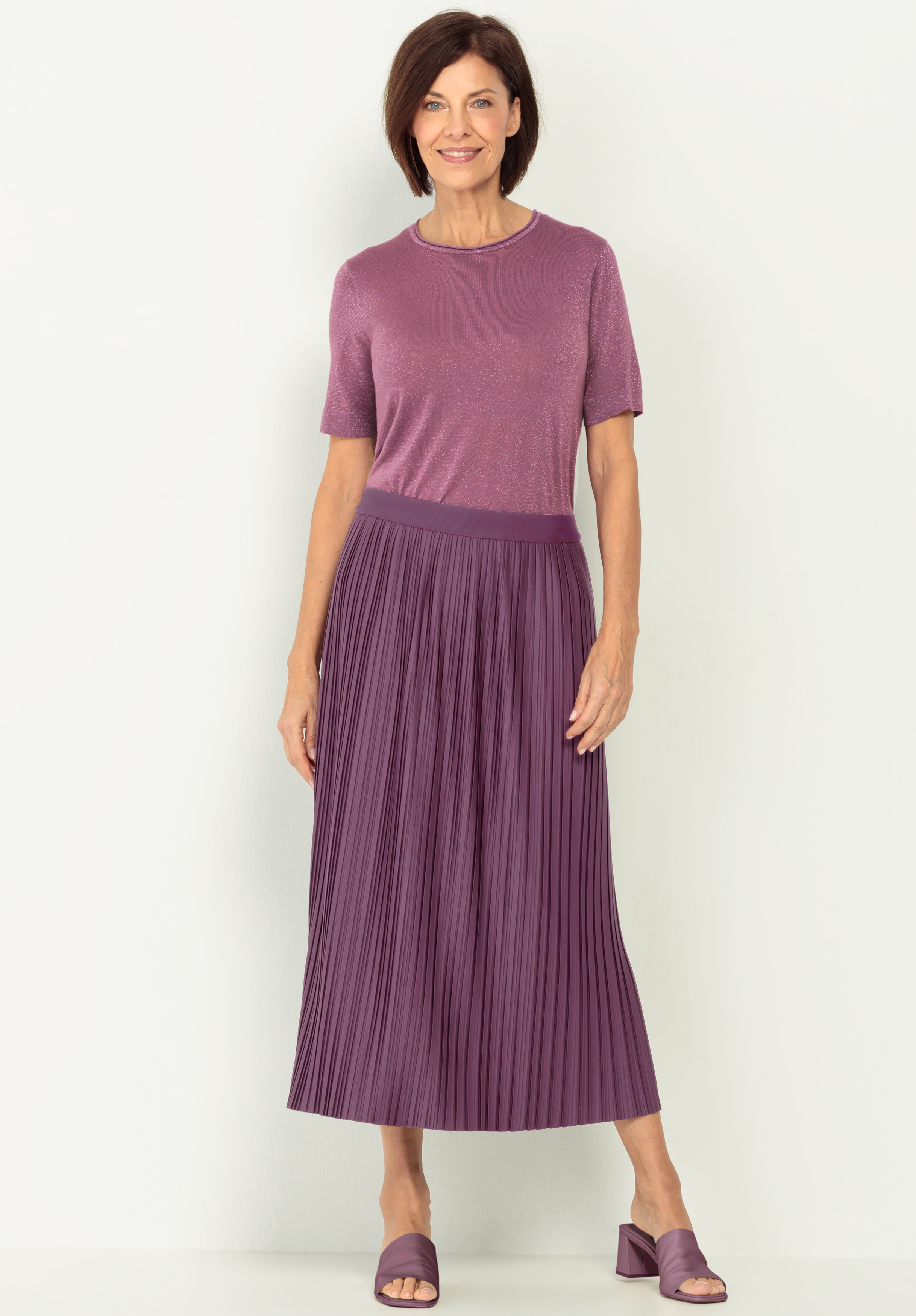 Eine Frau trägt ein kurzärmeliges, violettes Oberteil, das in einen hochtaillierten, gefalteten violetten Midirock gesteckt wird, und dazu passende violette hochhackige Sandalen. Sie steht vor einem schlichten hellen Hintergrund und lächelt sanft.