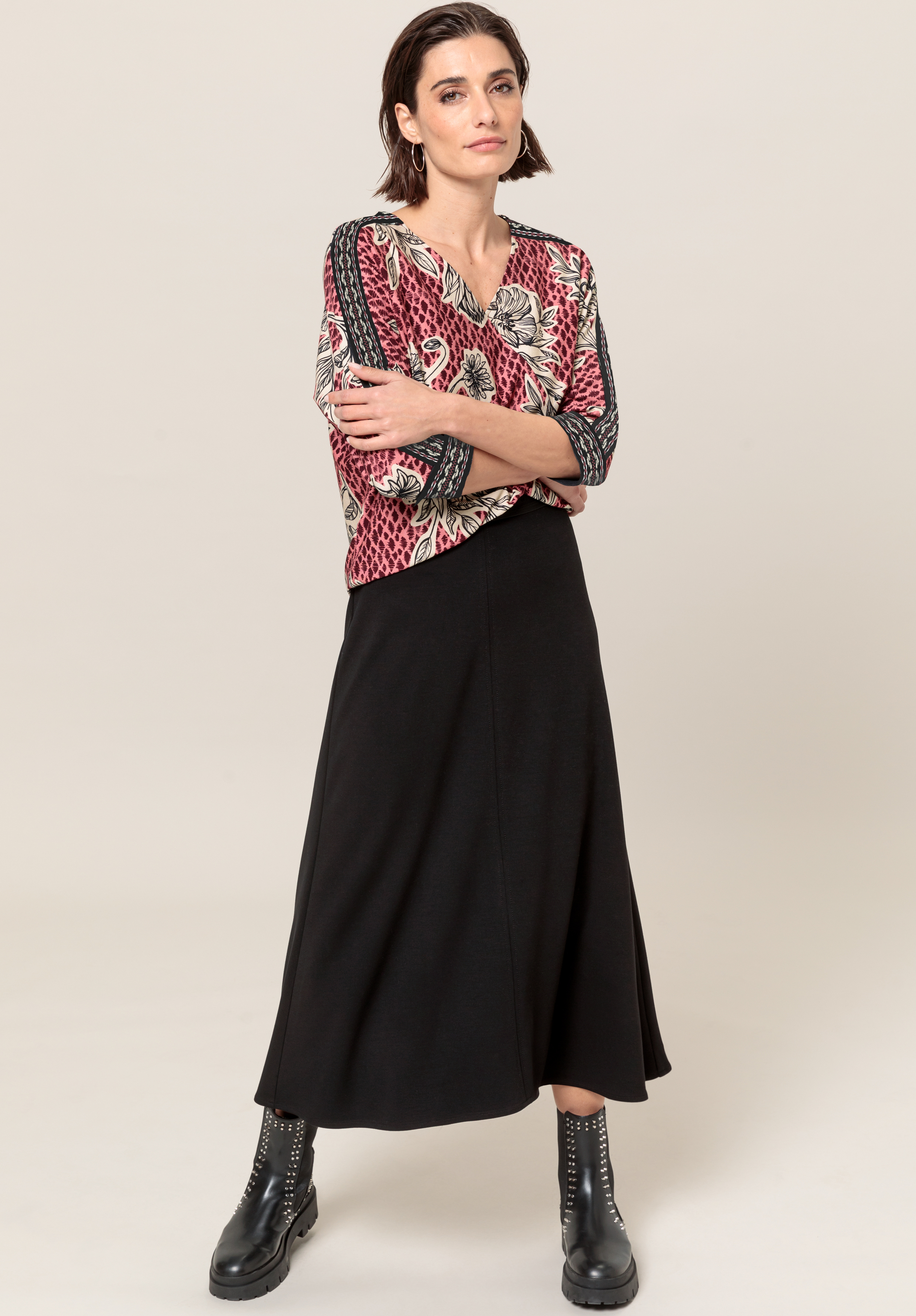 Eine Frau steht mit verschränkten Armen da und trägt ein gemustertes Langarmshirt, einen schwarzen A-Linien-Rock und schwarze Schnürstiefel. Sie hat kurzes dunkles Haar und einen neutralen Gesichtsausdruck vor einem schlichten Hintergrund.