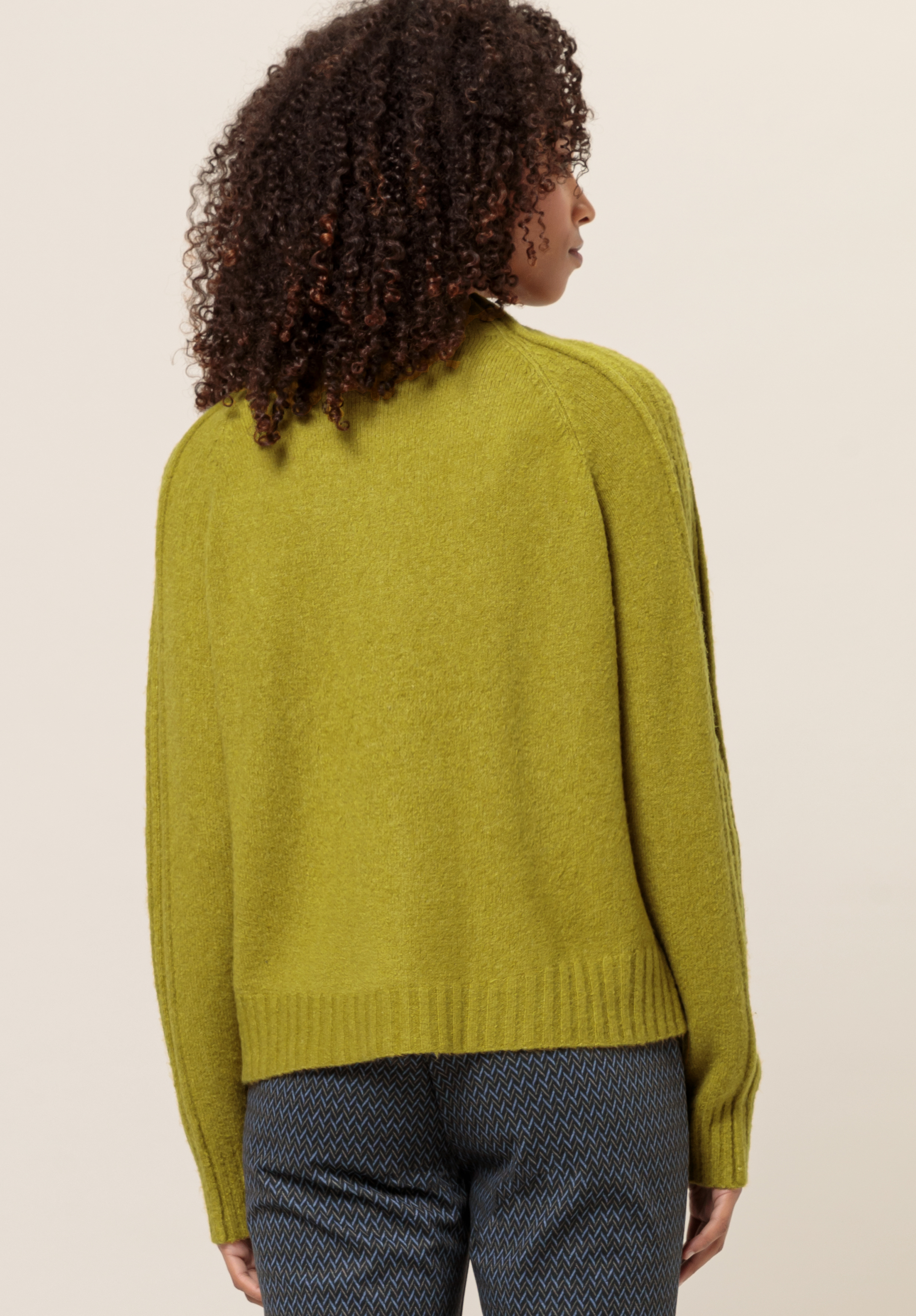 Von hinten ist eine Person mit lockigem Haar zu sehen, die vor einem schlichten Hintergrund einen locker sitzenden, langärmeligen, hellgrünen Pullover und eine hoch taillierte, blau gemusterte Hose trägt.