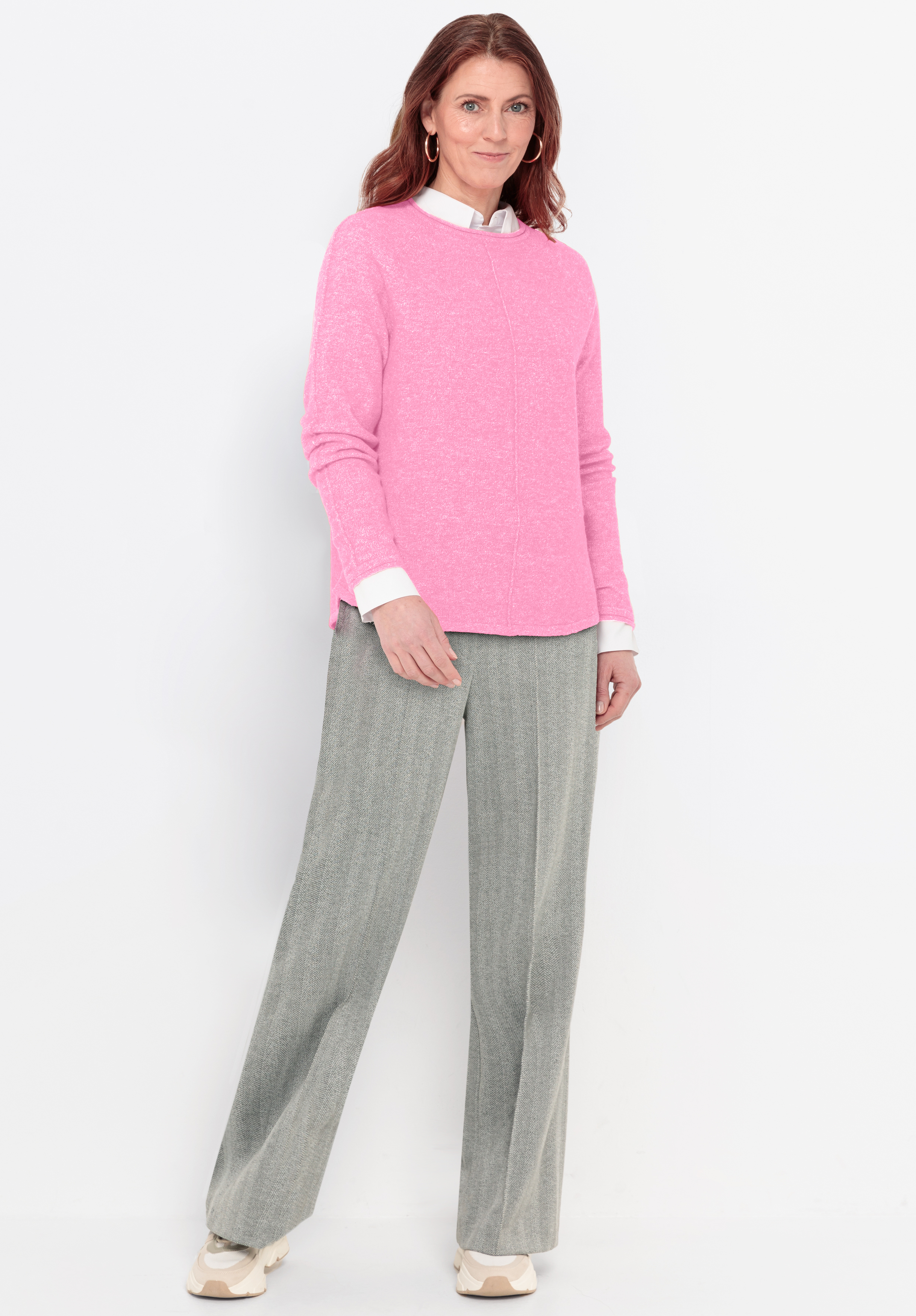 Eine Frau steht vor einem weißen Hintergrund und trägt einen hellrosa Pullover über einem weißen Hemd mit Kragen, eine graue Hose mit weitem Bein und weiße Turnschuhe. Sie hat schulterlanges kastanienbraunes Haar und lächelt leicht.