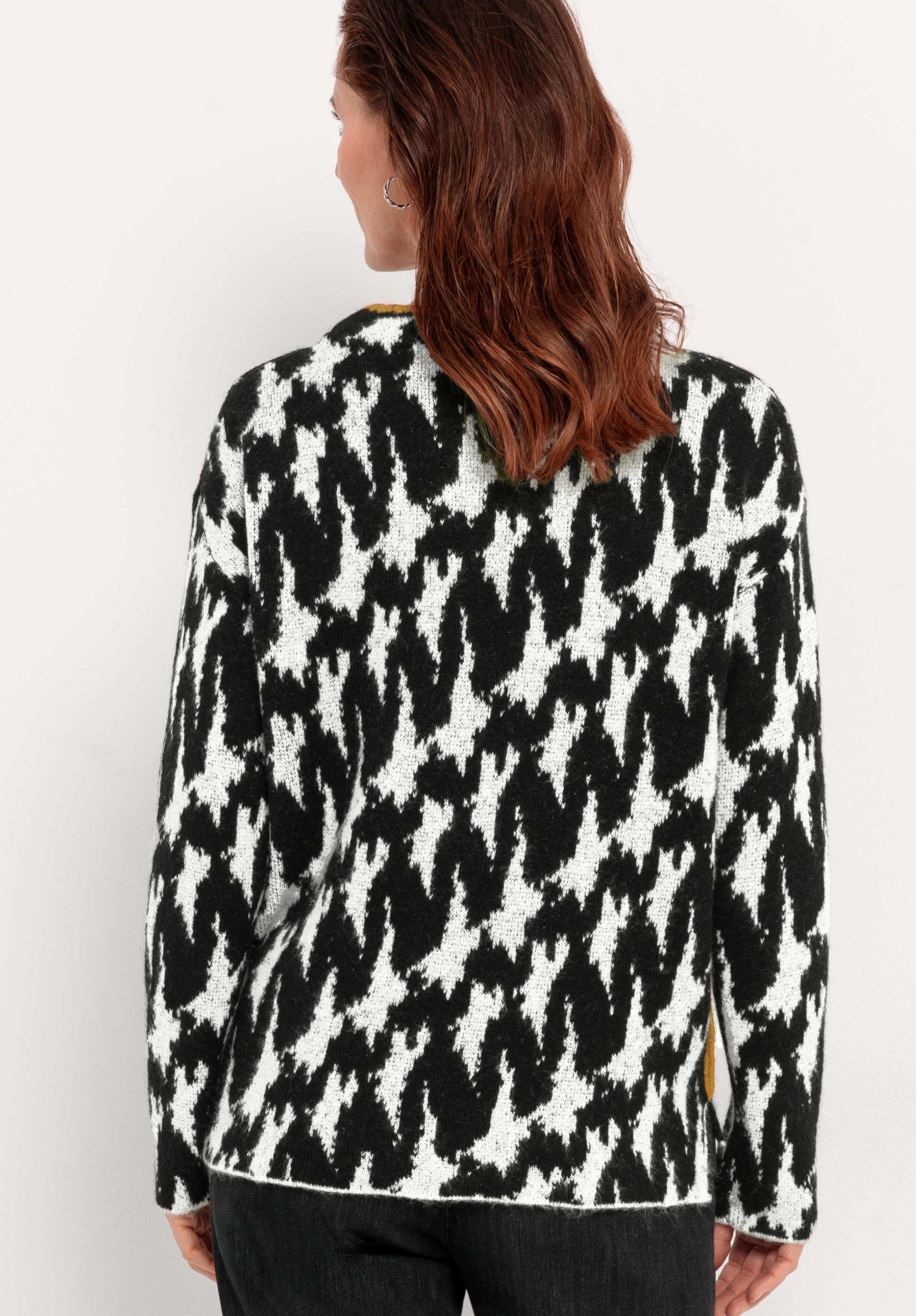 Eine Frau mit welligem braunem Haar ist von hinten zu sehen; sie trägt einen schwarz-weißen Pullover mit Hahnentrittmuster und eine dunkle Hose und steht vor einer schlichten, hellen Wand.