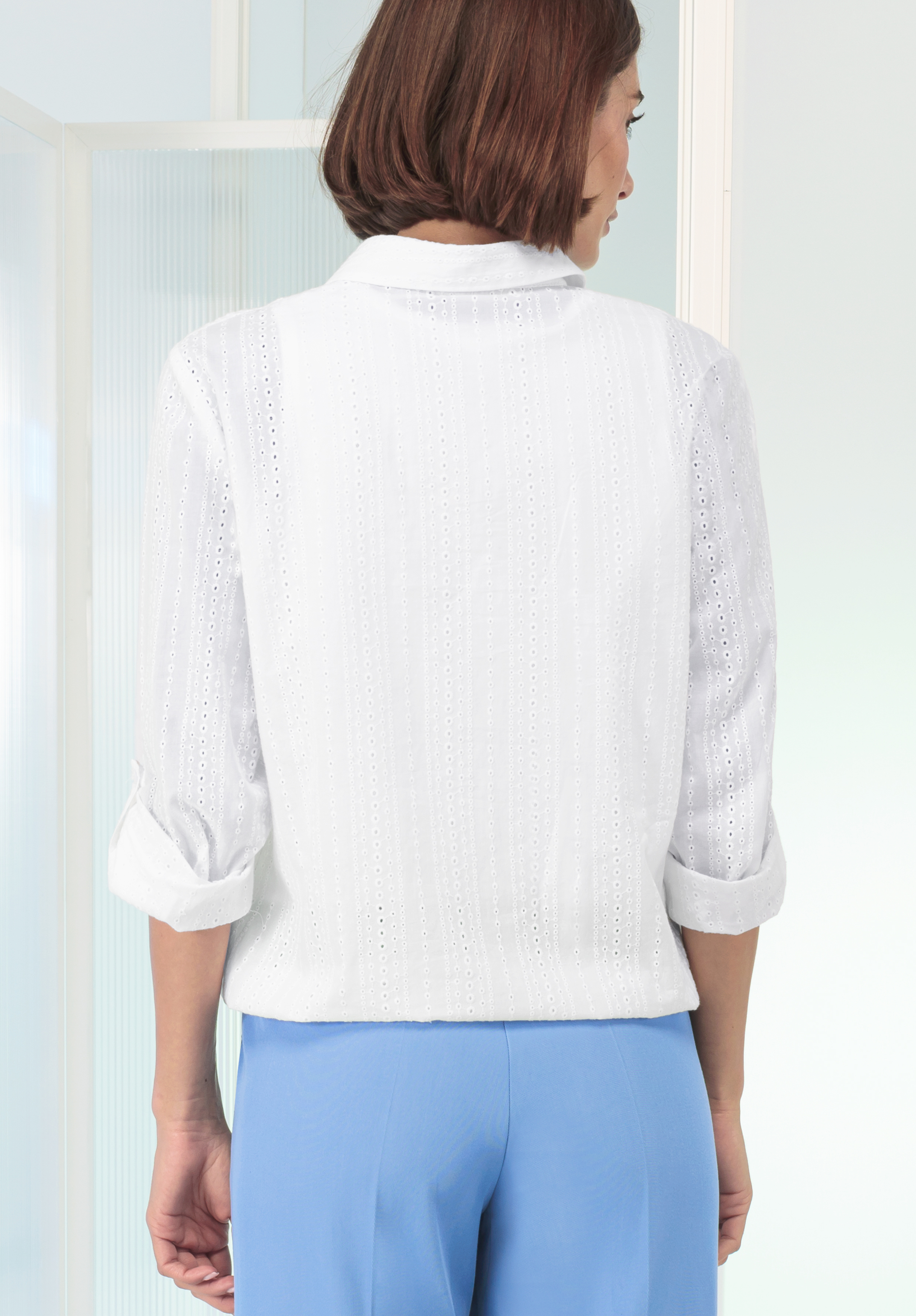 Eine Person mit kurzen braunen Haaren, von hinten gesehen, trägt ein weißes, strukturiertes Langarmhemd mit hochgekrempelten Ärmeln und hellblaue Hosen und steht in einer hellen, minimalistischen Innenumgebung.