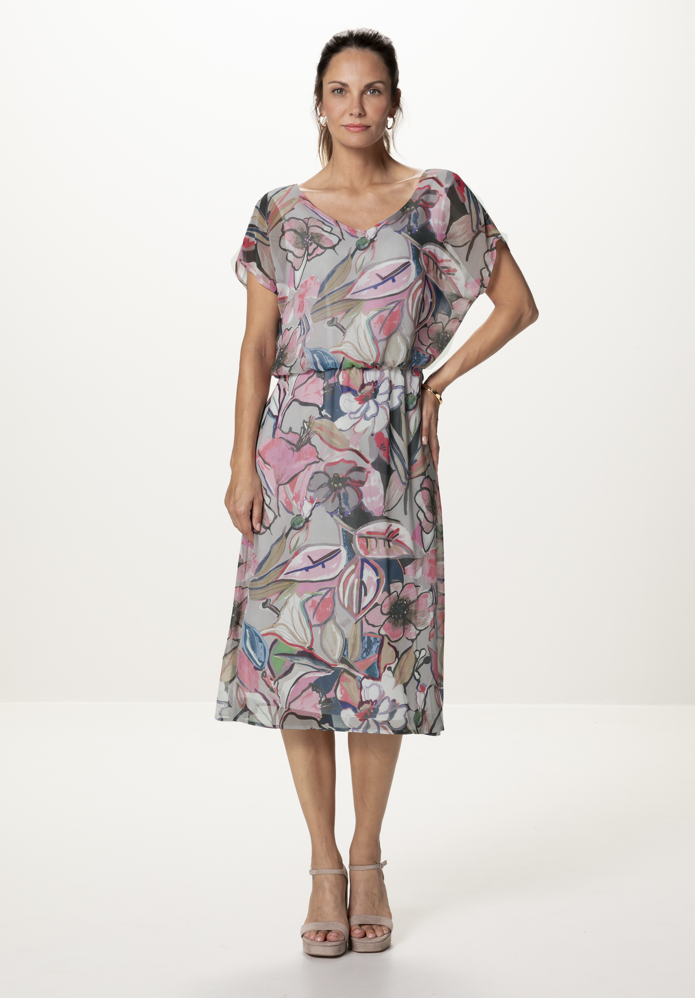Eine Frau steht in einem Studio. Sie trägt ein leichtes, knielanges Kleid mit Blumenmuster und kurzen Ärmeln sowie rosafarbene Absätze und posiert mit einer Hand auf der Hüfte vor einem schlichten weißen Hintergrund.
