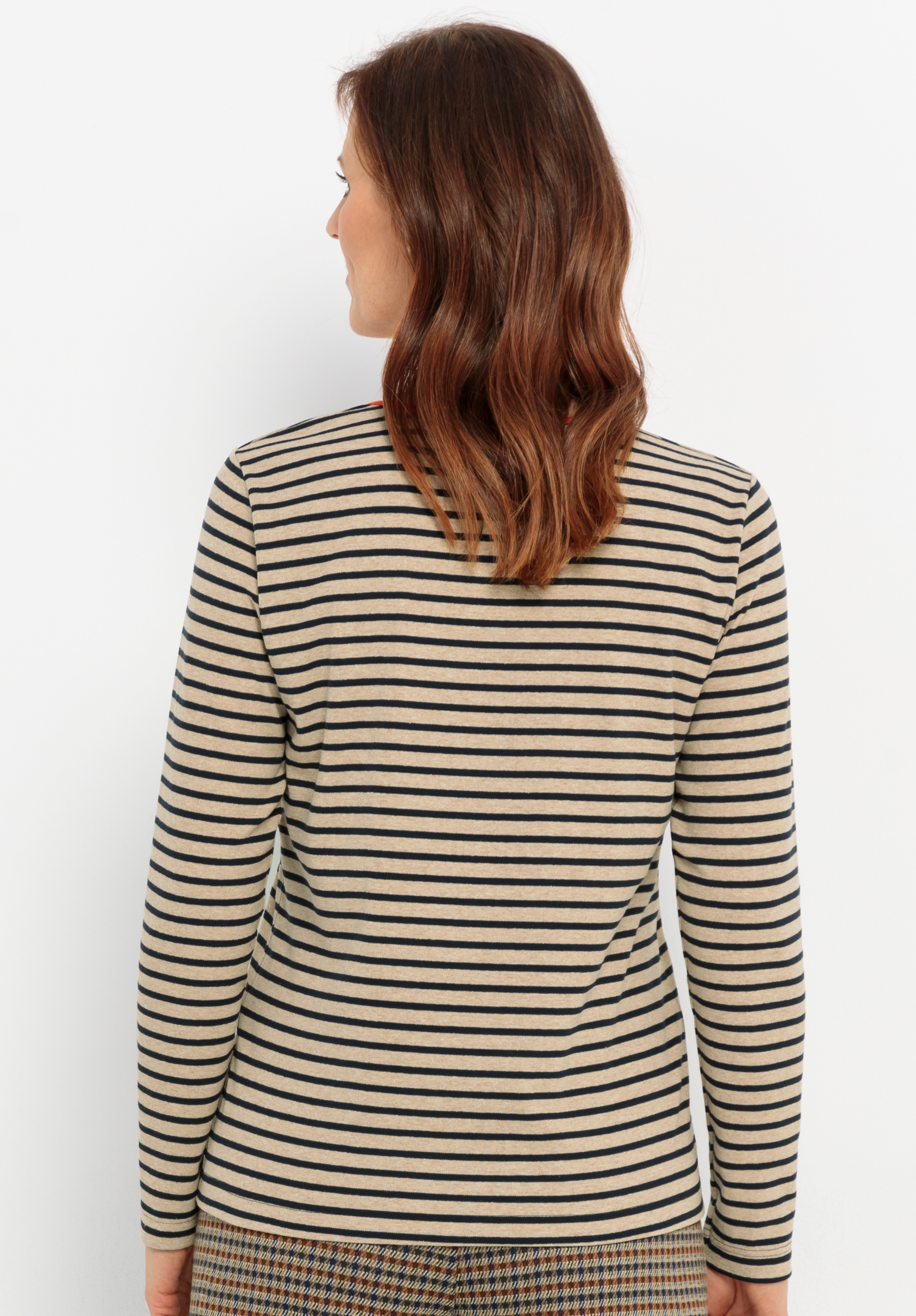 Eine Frau mit braunem, gewelltem Haar wird von hinten gezeigt. Sie trägt ein langärmeliges beiges Oberteil mit schwarzen Querstreifen und eine braun karierte Hose. Sie steht vor einem schlichten weißen Hintergrund.