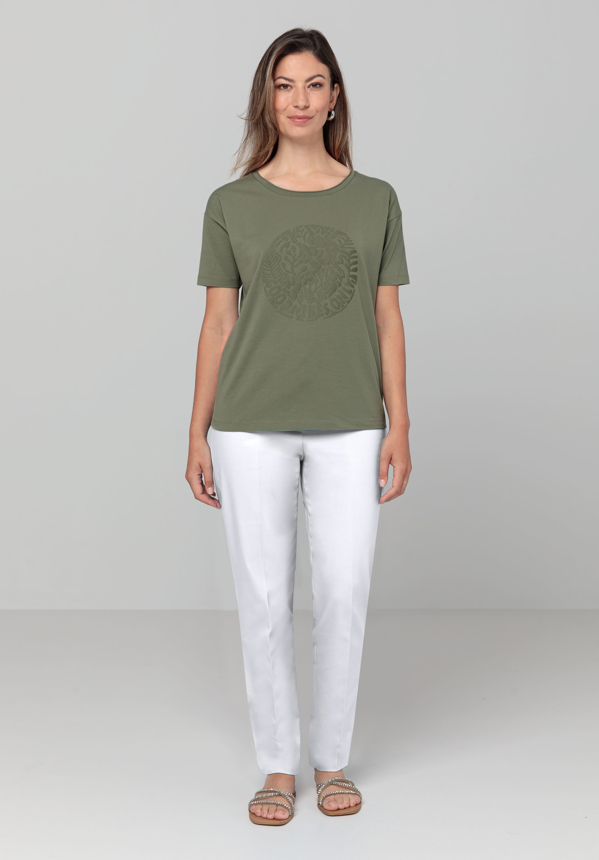 Eine Frau steht lächelnd nach vorne gerichtet und trägt ein salbeigrünes Kurzarm-T-Shirt mit dezentem Kreismuster und eine weiße Hose. Sie hat hellbraunes Haar und Sandalen. Der Hintergrund ist schlicht hellgrau.