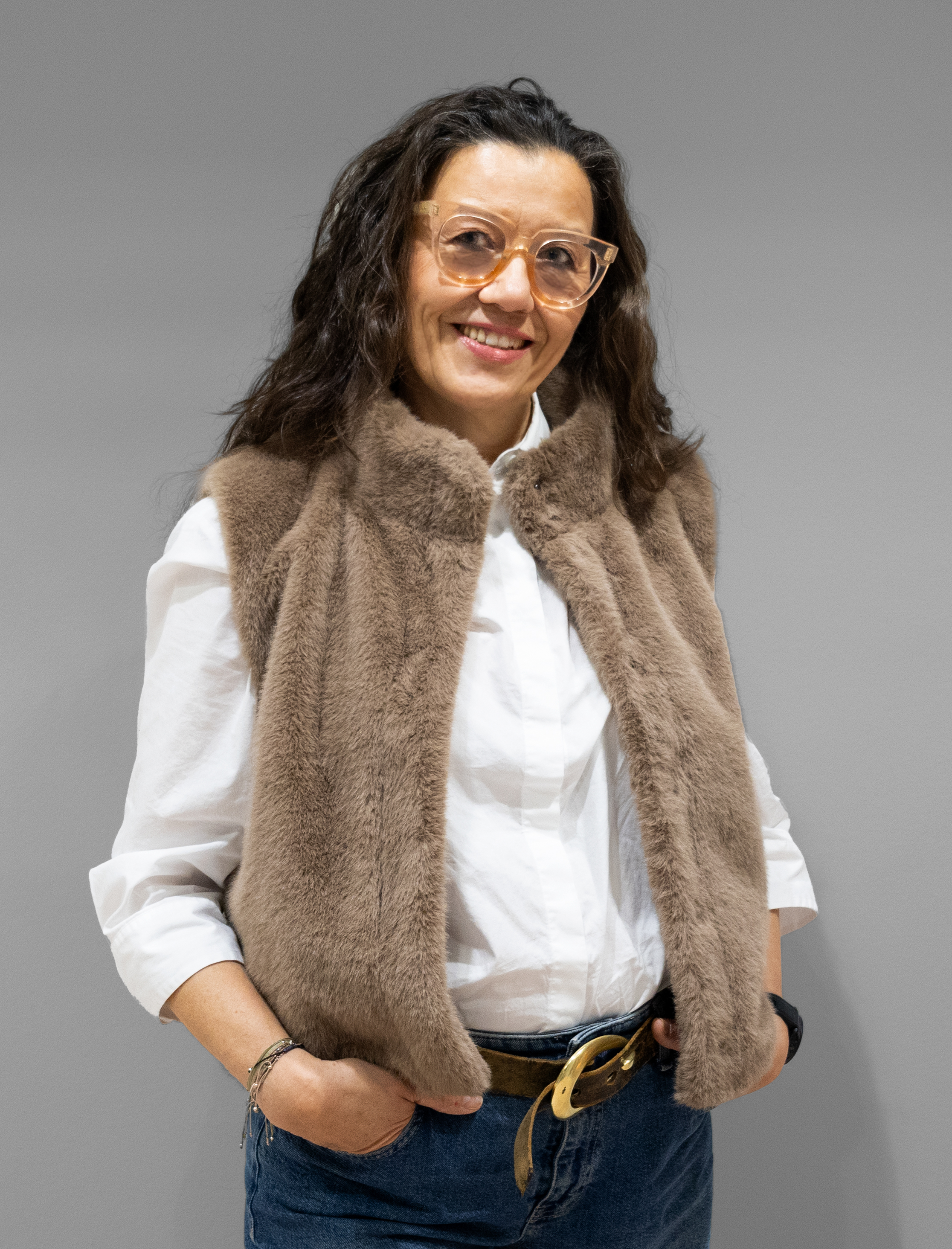 Eine Frau mit welligem braunem Haar und Brille lächelt in die Kamera. Sie trägt ein weißes Hemd, eine braune Kunstpelzweste und blaue Jeans und steht vor einem schlichten grauen Hintergrund.