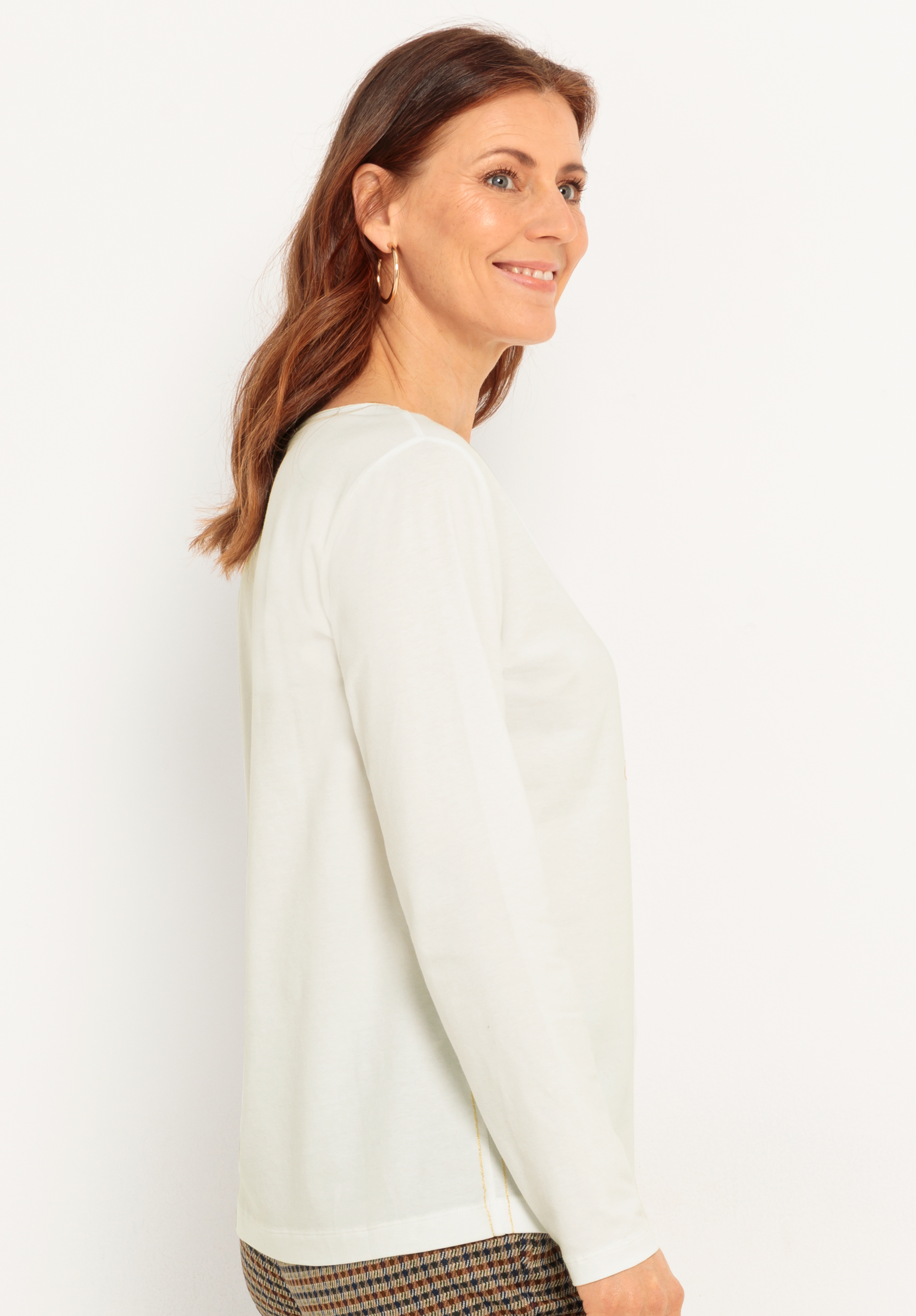 Eine Frau mit schulterlangen braunen Haaren, die ein langärmeliges weißes Oberteil und eine karierte Hose trägt, steht seitlich vor einem schlichten weißen Hintergrund, lächelt und blickt nach vorn.