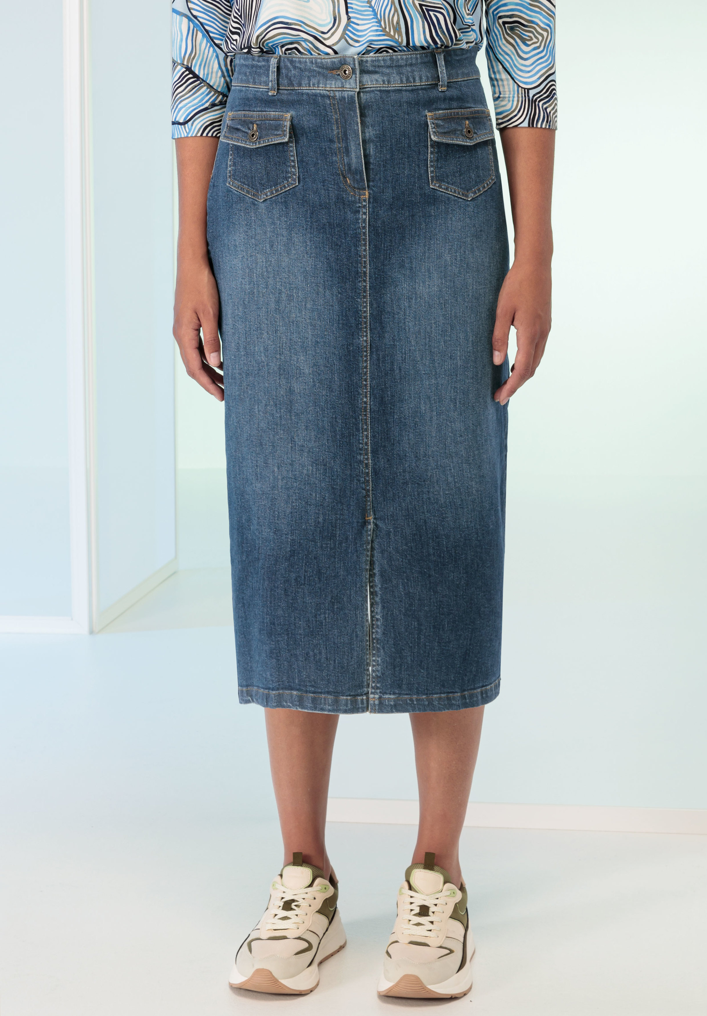 Jeansrock JONA Eine Person trägt einen blauen Jeans-Midirock mit Vordertaschen und beigen Turnschuhen und steht in einem minimalistischen, lichtdurchfluteten Raum. Nur die untere Körperhälfte ist sichtbar.