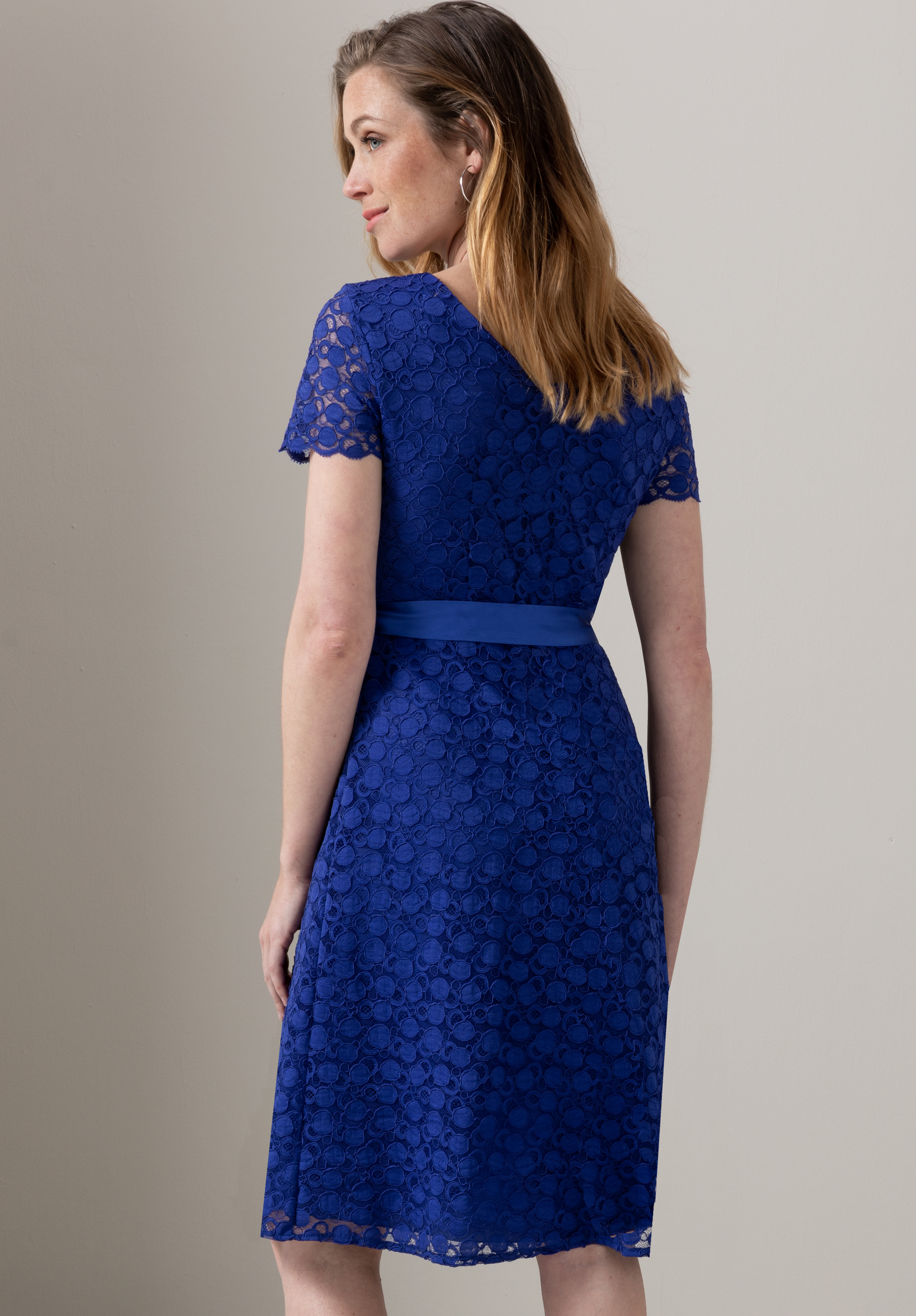 Eine Frau mit schulterlangem braunem Haar steht mit dem Rücken zur Kamera. Sie trägt ein knielanges, kurzärmeliges, königsblaues Spitzenkleid mit passendem Taillenbund. Sie befindet sich vor einer schlichten, hellen Wand.