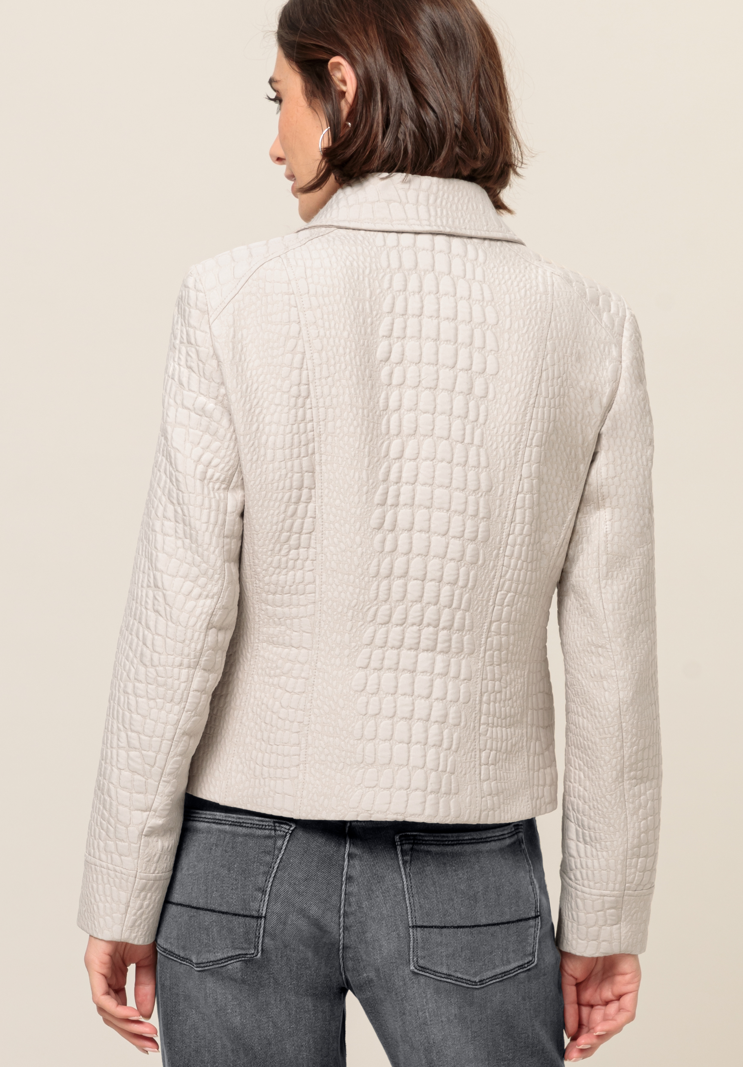 Von hinten ist eine Frau mit kurzen braunen Haaren zu sehen, die vor einem schlichten Hintergrund eine hellbeige, strukturierte Jacke mit Krokodilmuster und dunkelgraue Jeans mit sichtbaren Gesäßtaschen trägt.
