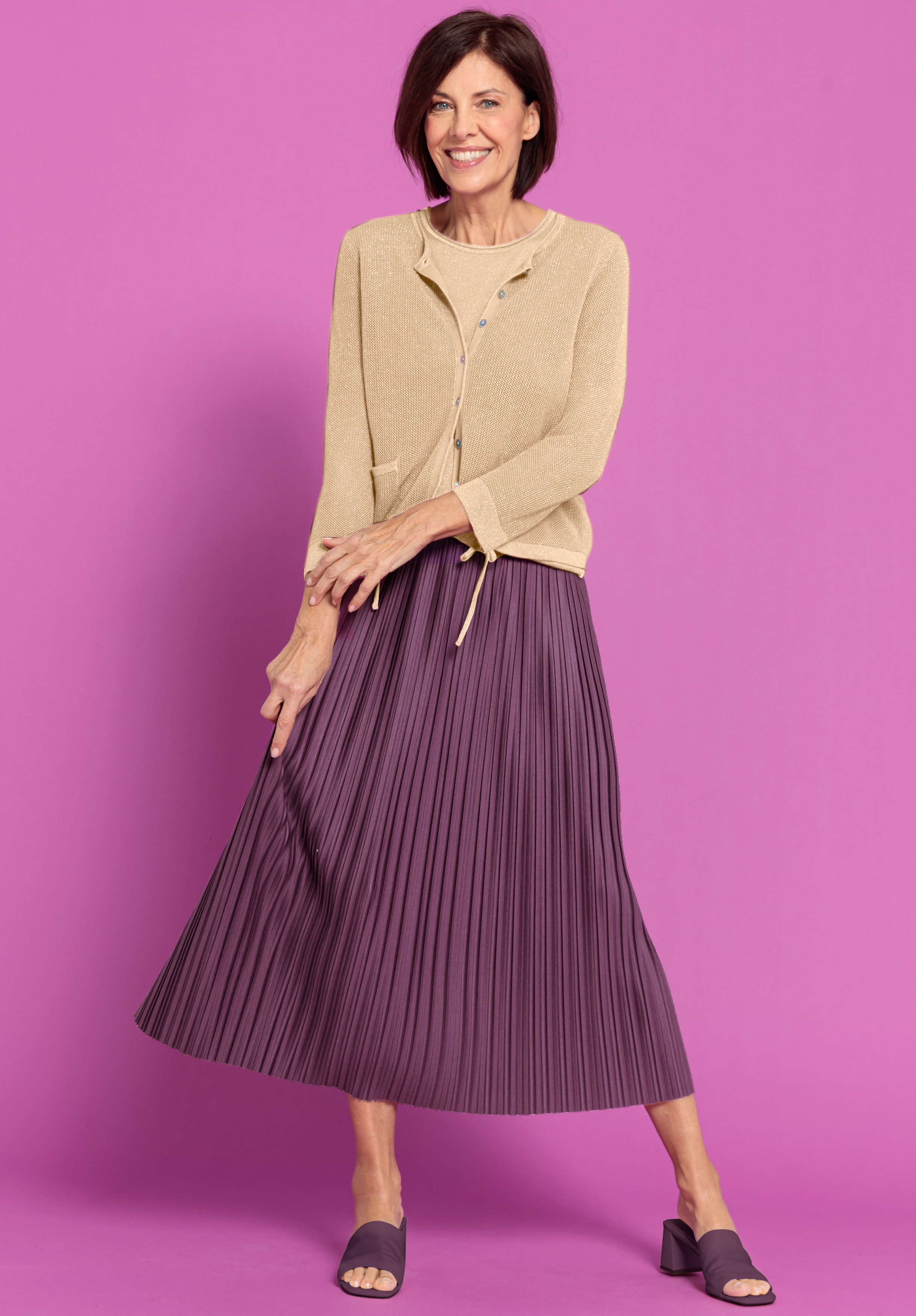 Eine Frau steht vor einem rosafarbenen Hintergrund, lächelt und trägt eine beigefarbene Strickjacke, einen plissierten lila Midirock und passende lila offene Schuhe.