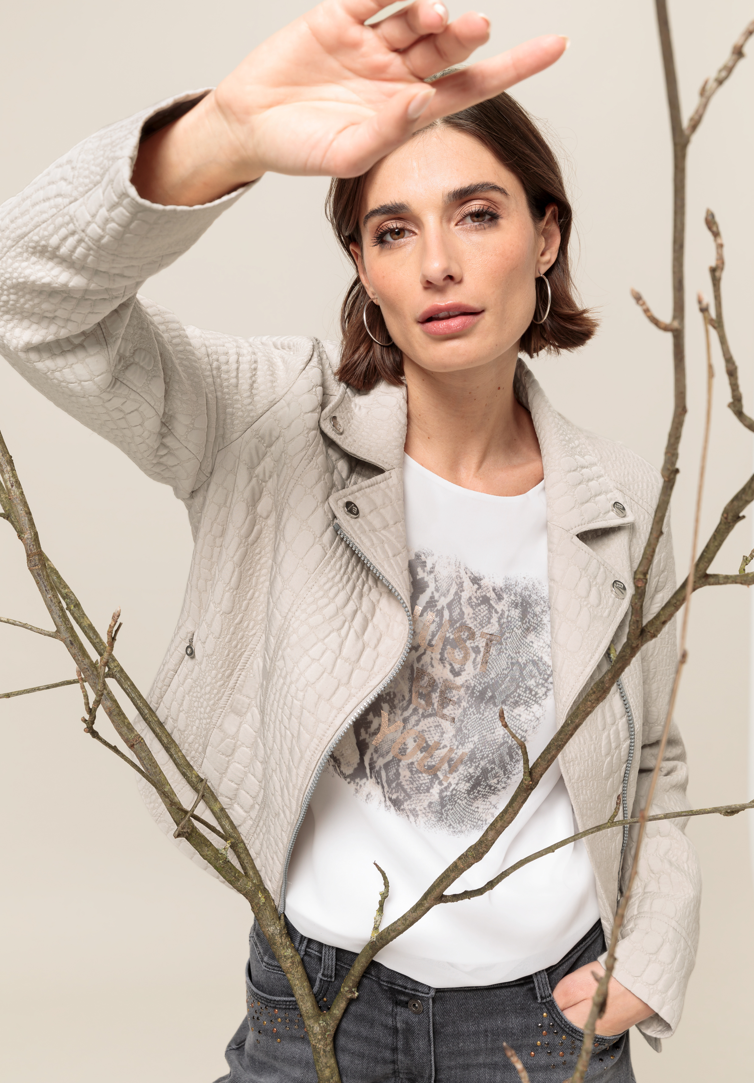 Bikerjacke SILJA Eine Frau in einer hellen Strukturjacke und einem T-Shirt mit Aufdruck steht hinter kahlen Ästen und hält sich die Hand vors Gesicht. Sie hat kurzes braunes Haar, Creolen und einen neutralen Gesichtsausdruck.