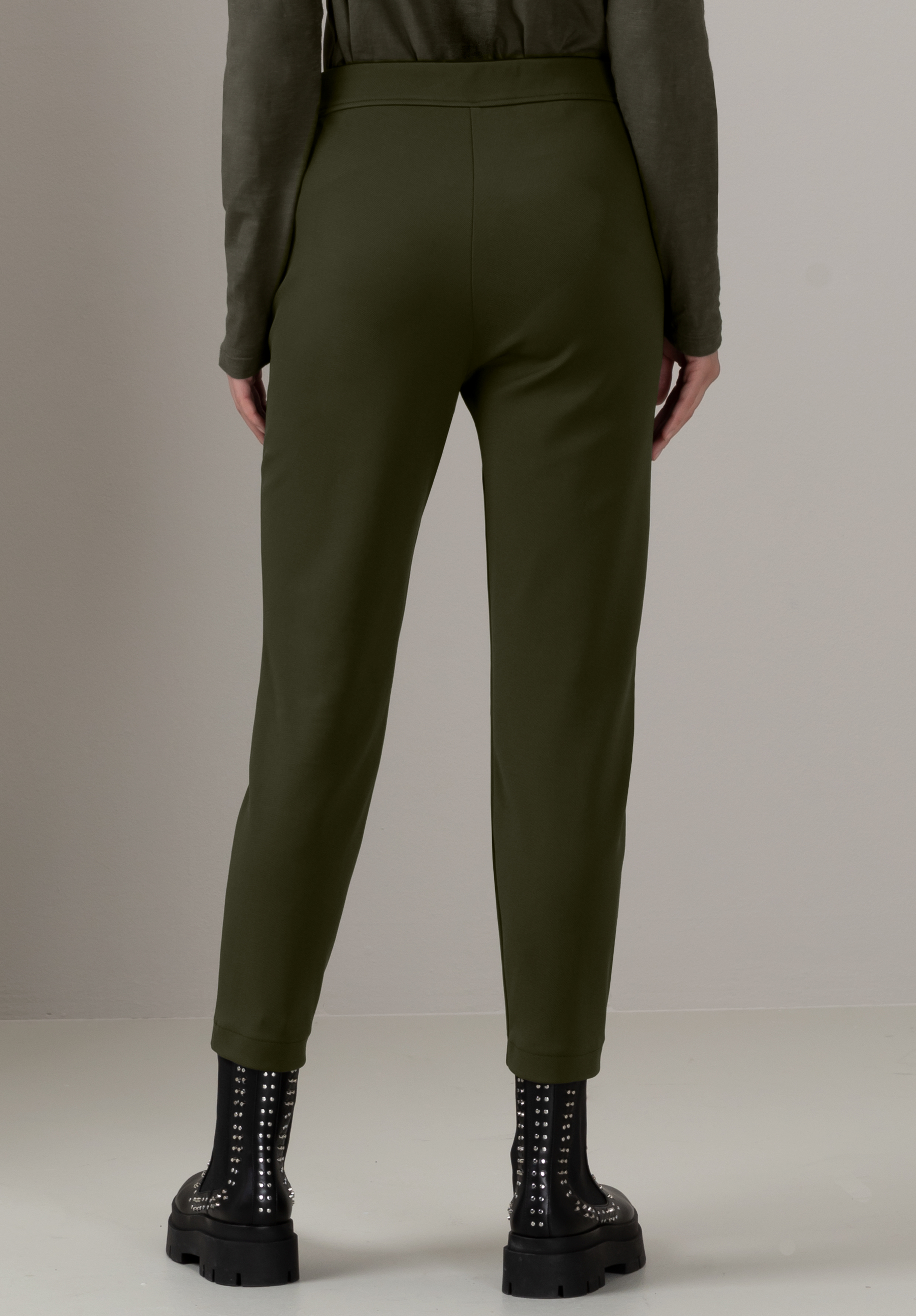 Eine Person steht mit abgewandtem Gesicht da und trägt hoch taillierte olivgrüne Hosen und schwarze Stiefeletten mit silbernen Nieten vor einem schlichten hellen Hintergrund.