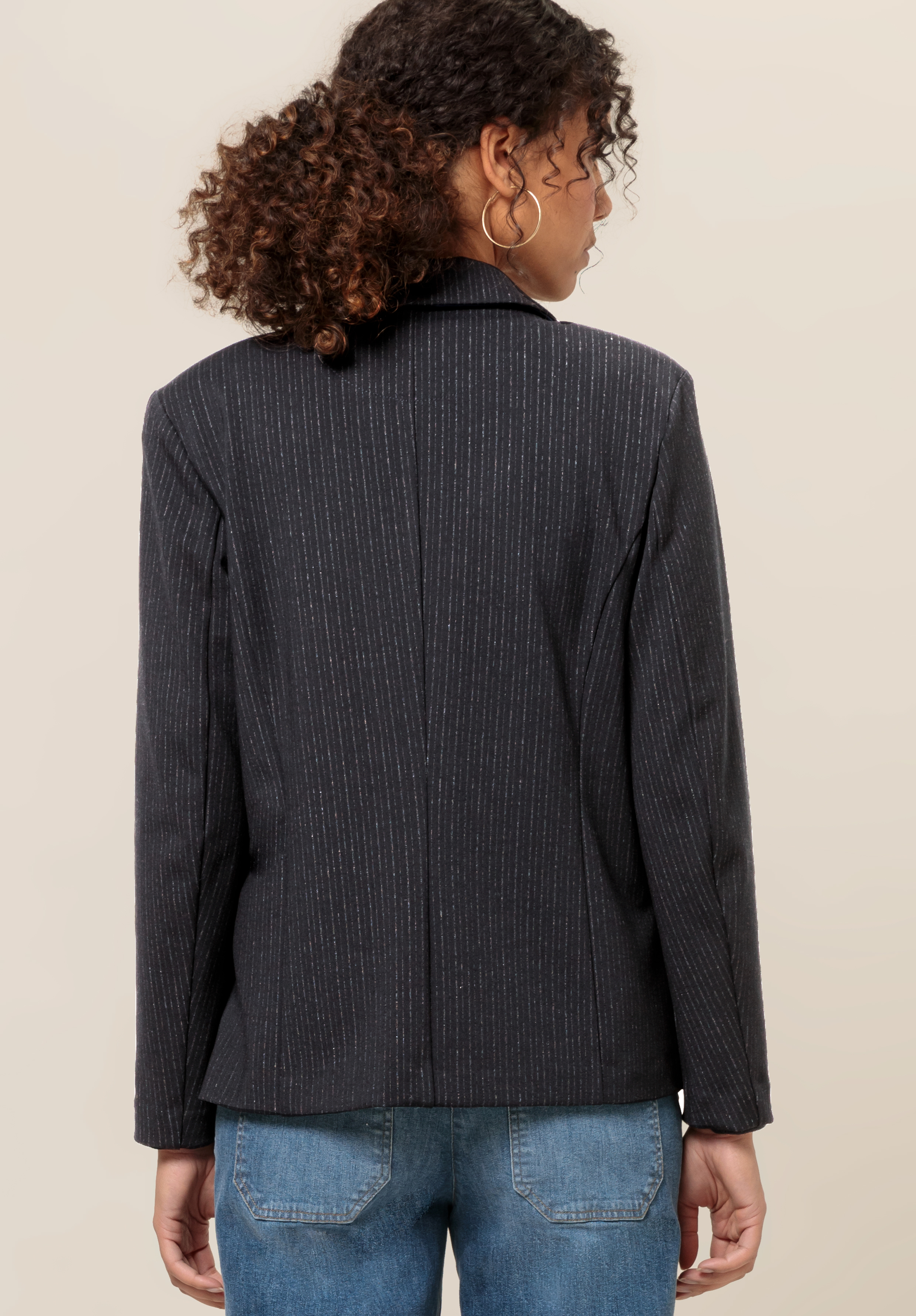 Eine Person mit lockigem Haar, Creolen, einem dunklen Nadelstreifenblazer und Bluejeans steht mit dem Rücken zur Kamera vor einem schlichten beigen Hintergrund.