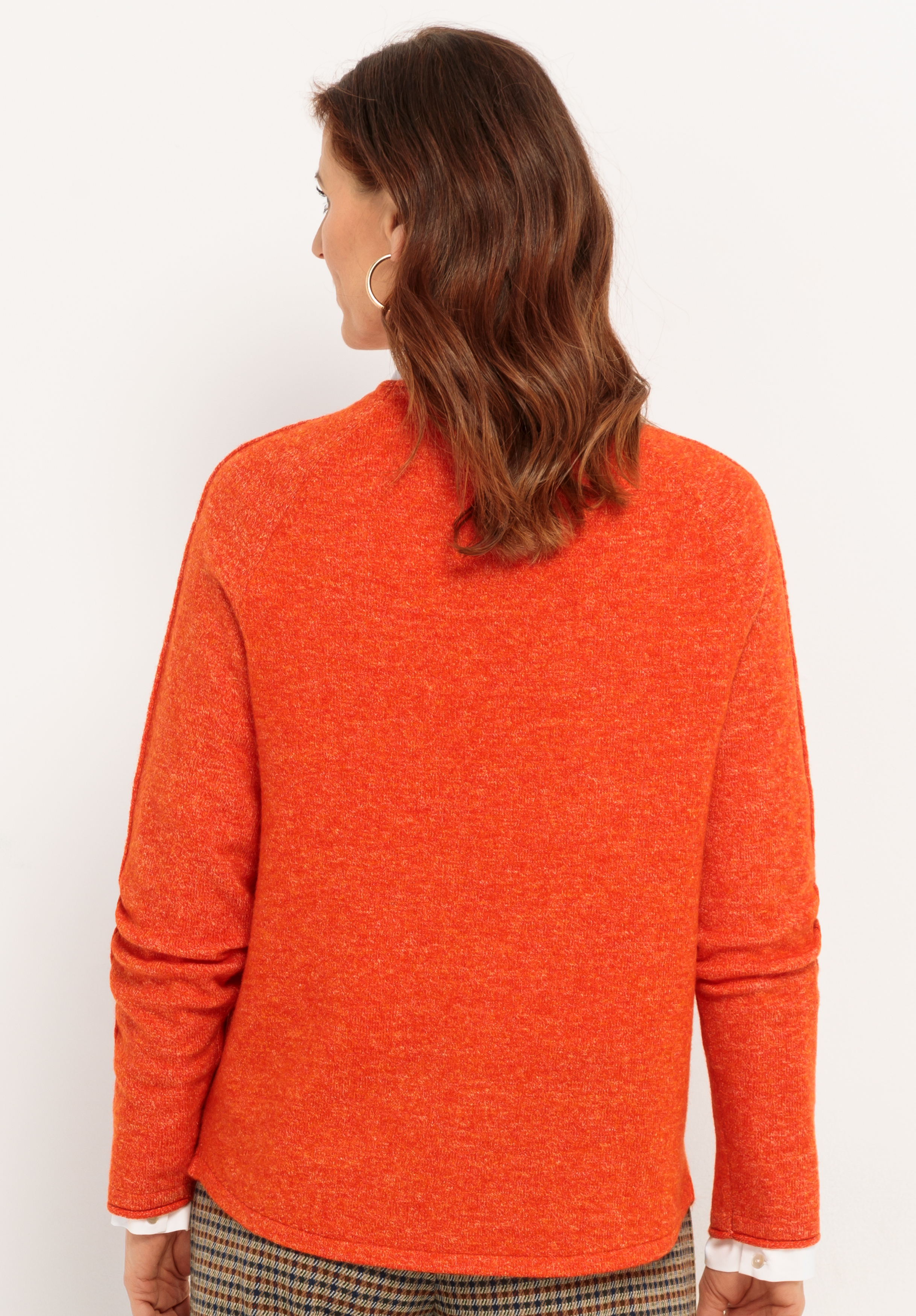 Eine Frau mit gewelltem braunem Haar, von hinten gesehen, trägt einen orangefarbenen Pullover und eine karierte Hose. Sie trägt Ohrringe und ein weißes Hemd, das an den Manschetten herausschaut. Der Hintergrund ist einfarbig weiß.