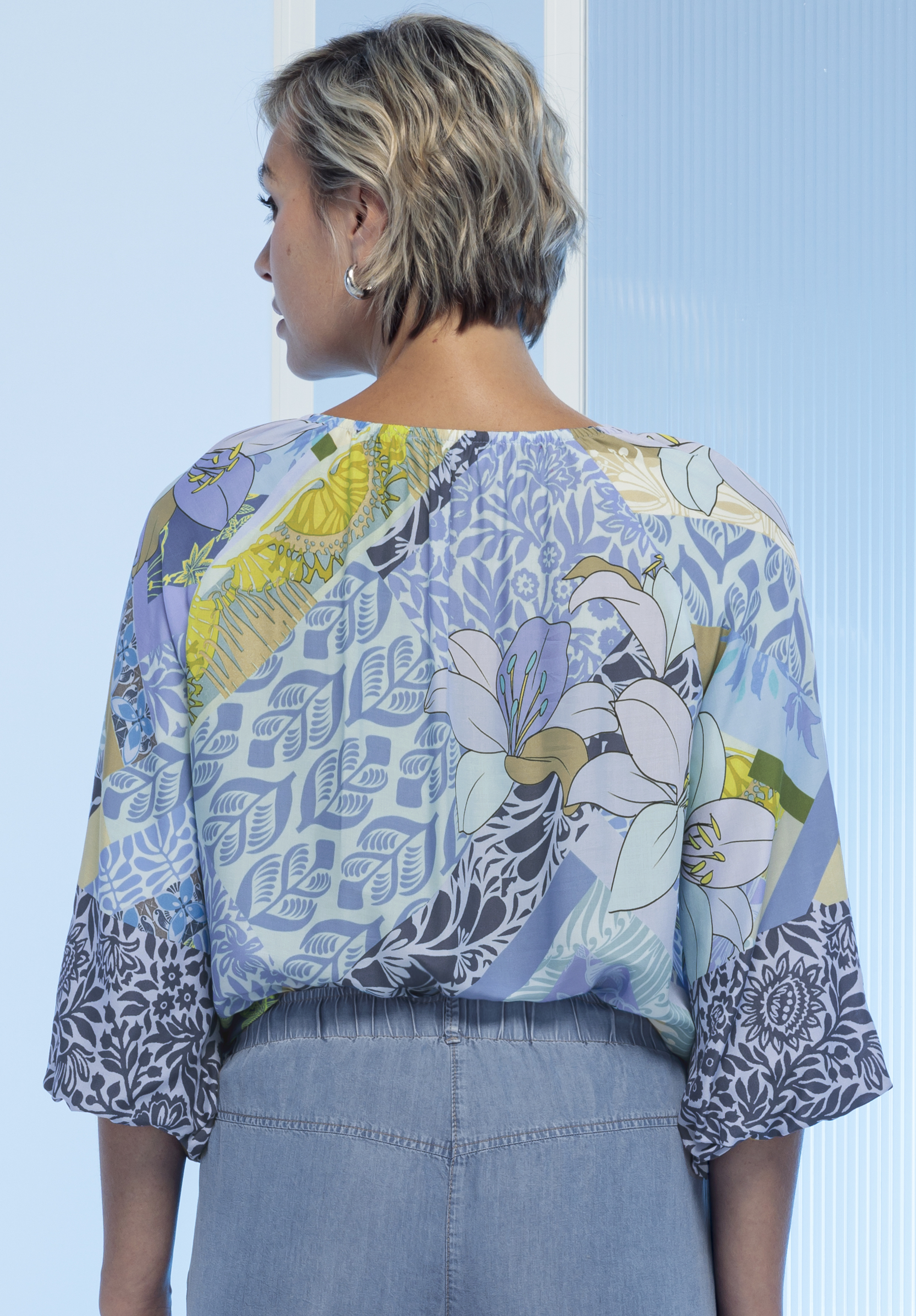 Eine Person mit kurzem, gewelltem Haar ist von hinten zu sehen. Sie trägt eine bunte, blumengemusterte Bluse mit weiten Ärmeln und eine hellblaue Hose mit hoher Taille. Der Hintergrund ist sanft in blassen Blautönen beleuchtet.