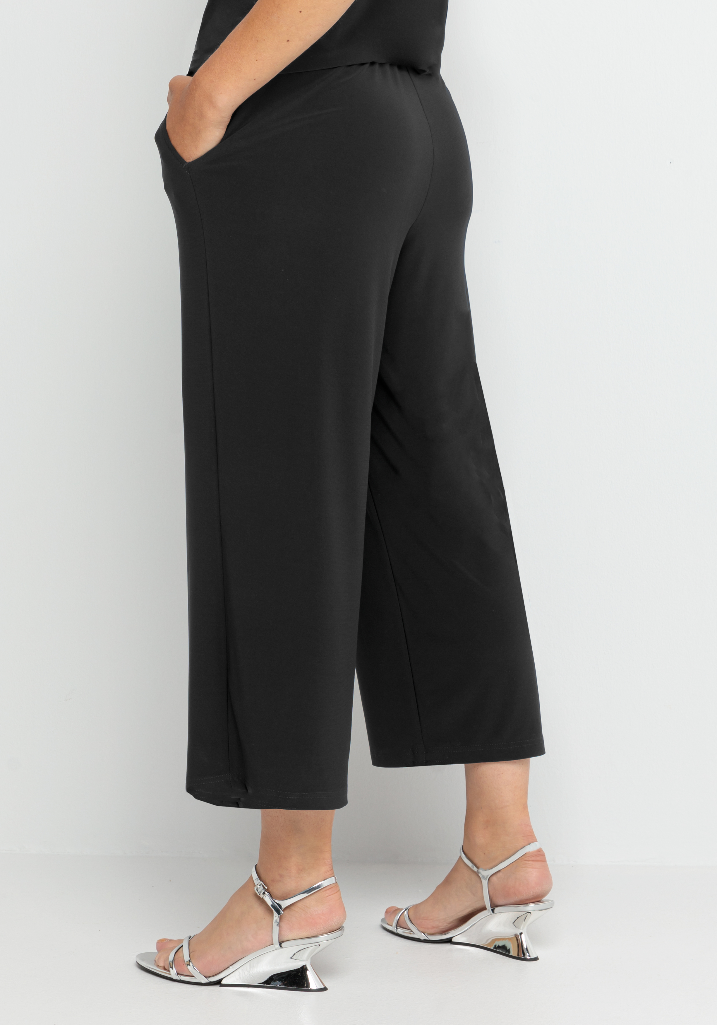 Eine Person, die eine schwarze Hose mit weitem Bein und silberne Sandalen mit Absätzen trägt, steht vor einem schlichten weißen Hintergrund und hat eine Hand in der Tasche.