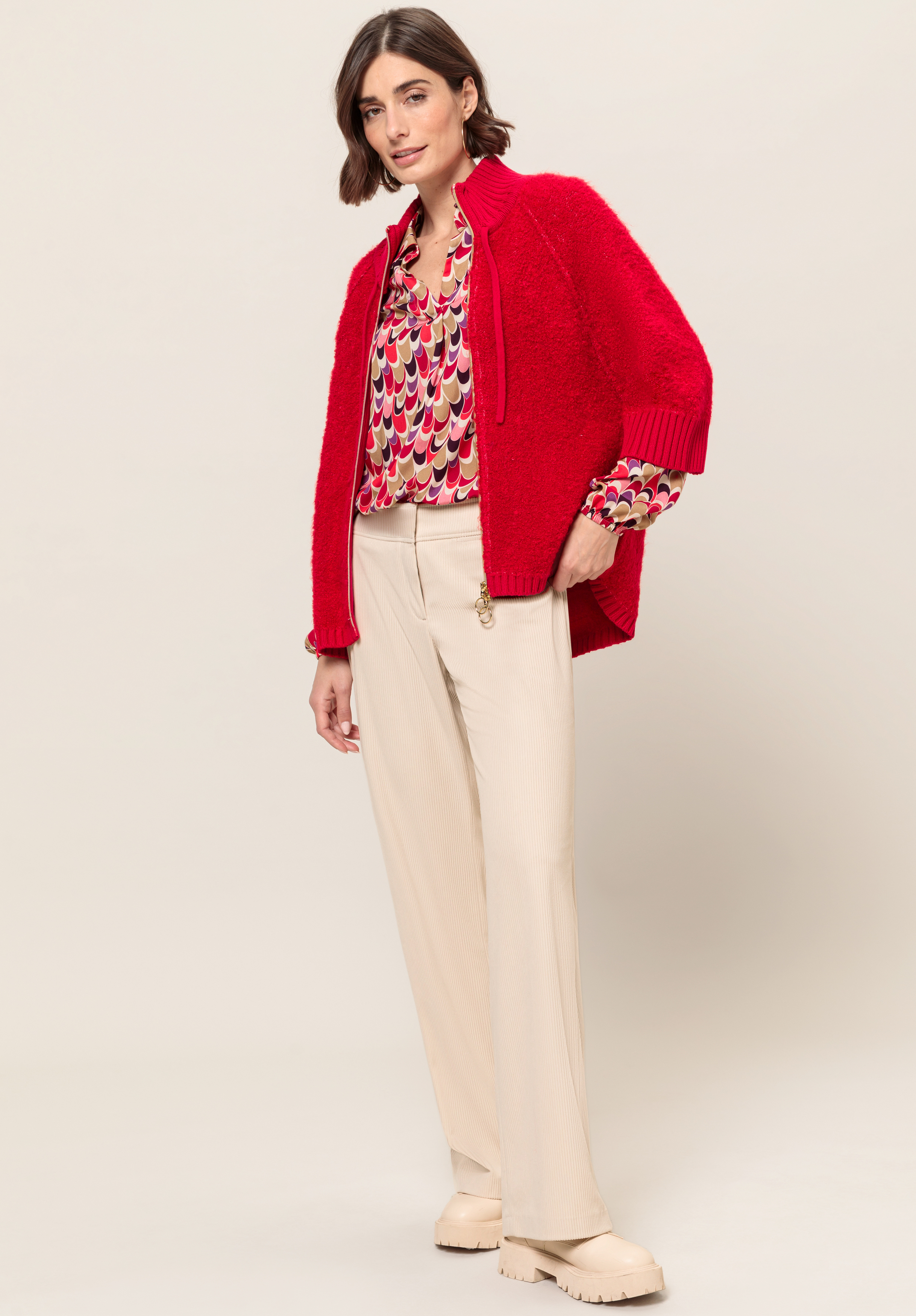 Vor einem schlichten Hintergrund steht eine Frau mit einer leuchtend roten Strickjacke, einer gemusterten Bluse mit roten, rosa und beigen Formen, beigen Hosen mit geradem Bein und klobigen cremefarbenen Schuhen.