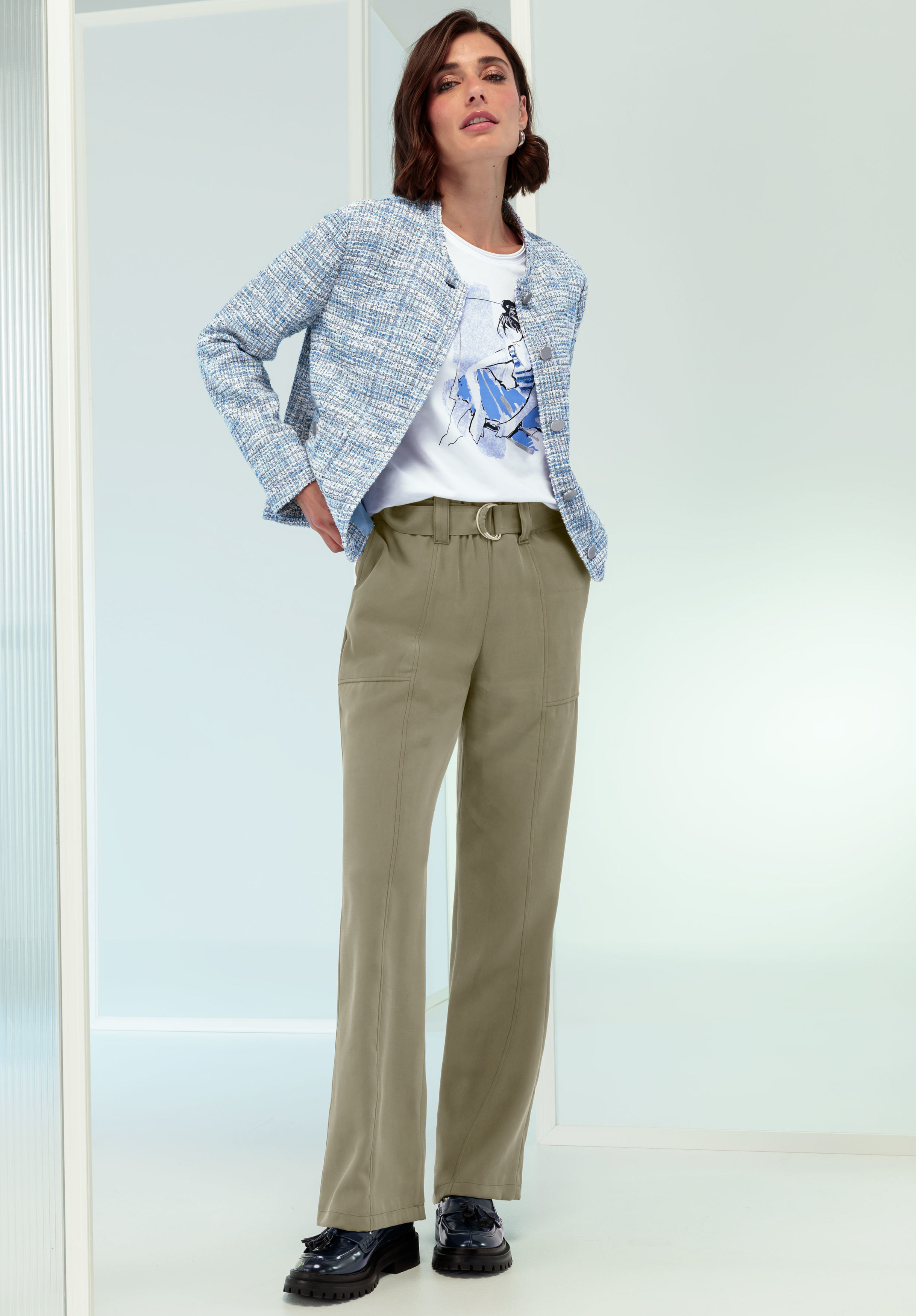 Eine Frau steht drinnen und trägt eine hellblaue Strukturjacke, ein weißes T-Shirt mit Aufdruck, khakifarbene Hosen mit hoher Taille und Gürtel sowie schwarze Schuhe. Ihre Hände stecken in den Taschen.