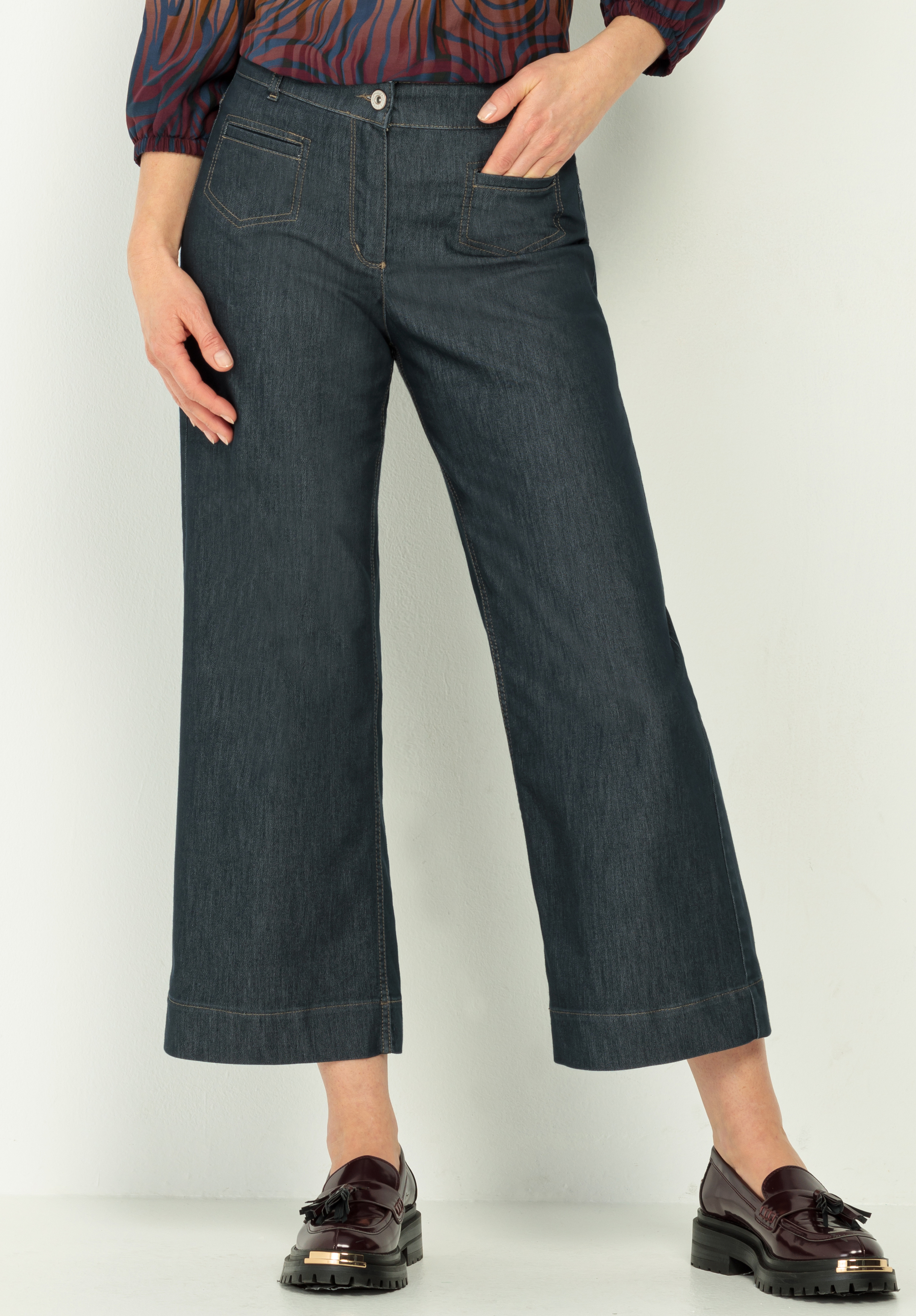 Stretch-Hose MELBOURNE Eine Person, die dunkle Jeans mit weitem Bein, ein gemustertes langärmeliges Oberteil und dunkle Slipper mit Quasten trägt, steht vor einem hellen Hintergrund.