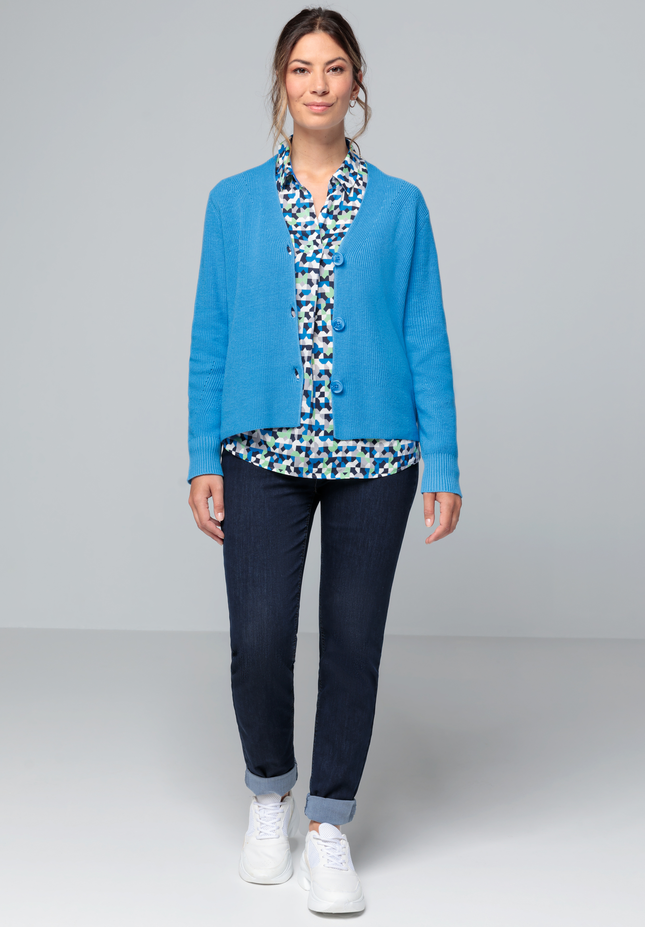 Eine Frau steht lächelnd nach vorne gerichtet. Sie trägt eine hellblaue Strickjacke über einer blau gemusterten Bluse, dunkle Jeans mit hochgekrempelten Hosenbeinen und weiße Turnschuhe. Der Hintergrund ist schlicht hellgrau.