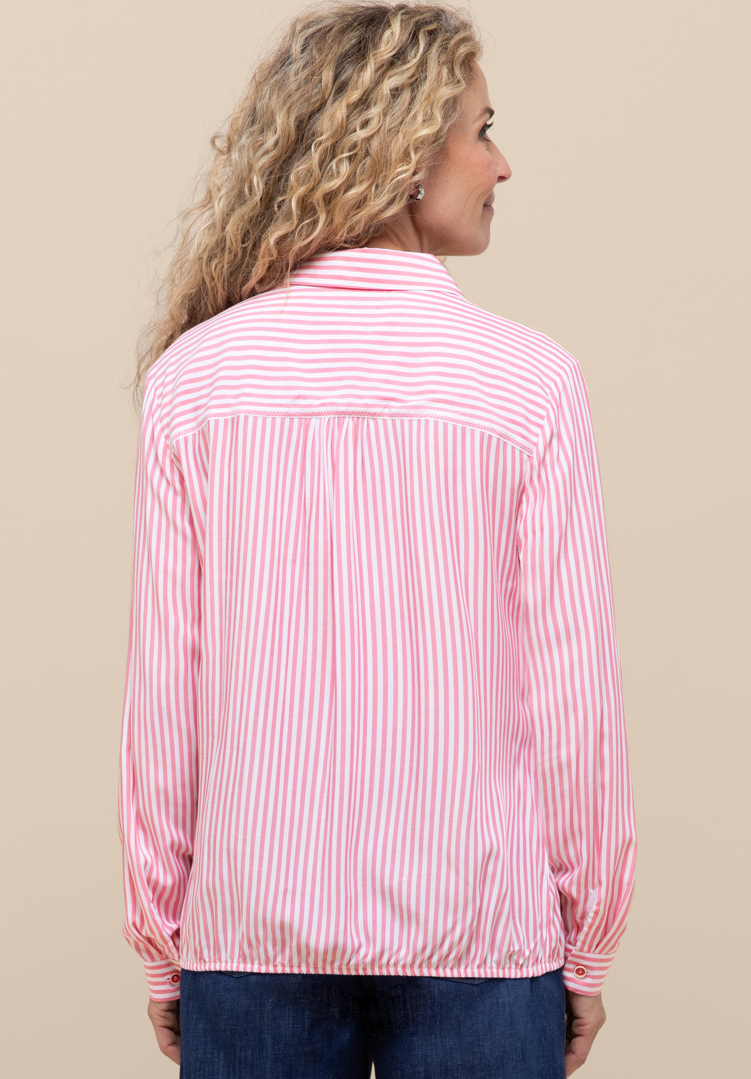Eine Frau mit langen, lockigen blonden Haaren steht mit dem Rücken zur Kamera, trägt eine rosa-weiß vertikal gestreifte Bluse und blaue Jeans und steht vor einem beigen Hintergrund.