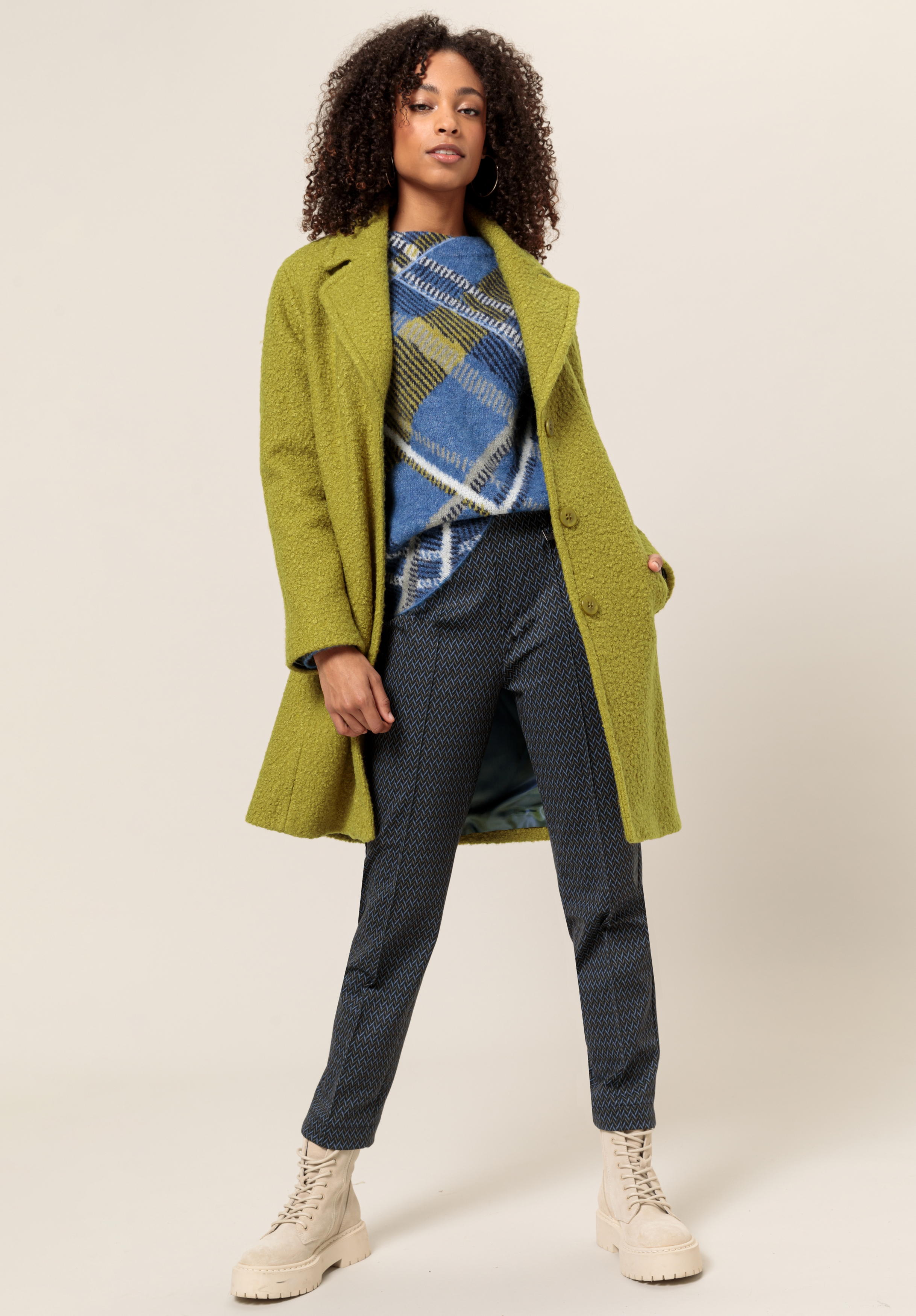 Eine Frau mit lockigem Haar trägt einen strukturierten grünen Mantel über einem blau gemusterten Pullover und einer dunkel gemusterten Hose, kombiniert mit hellen Schnürstiefeln, und steht selbstbewusst vor einem neutralen Hintergrund.