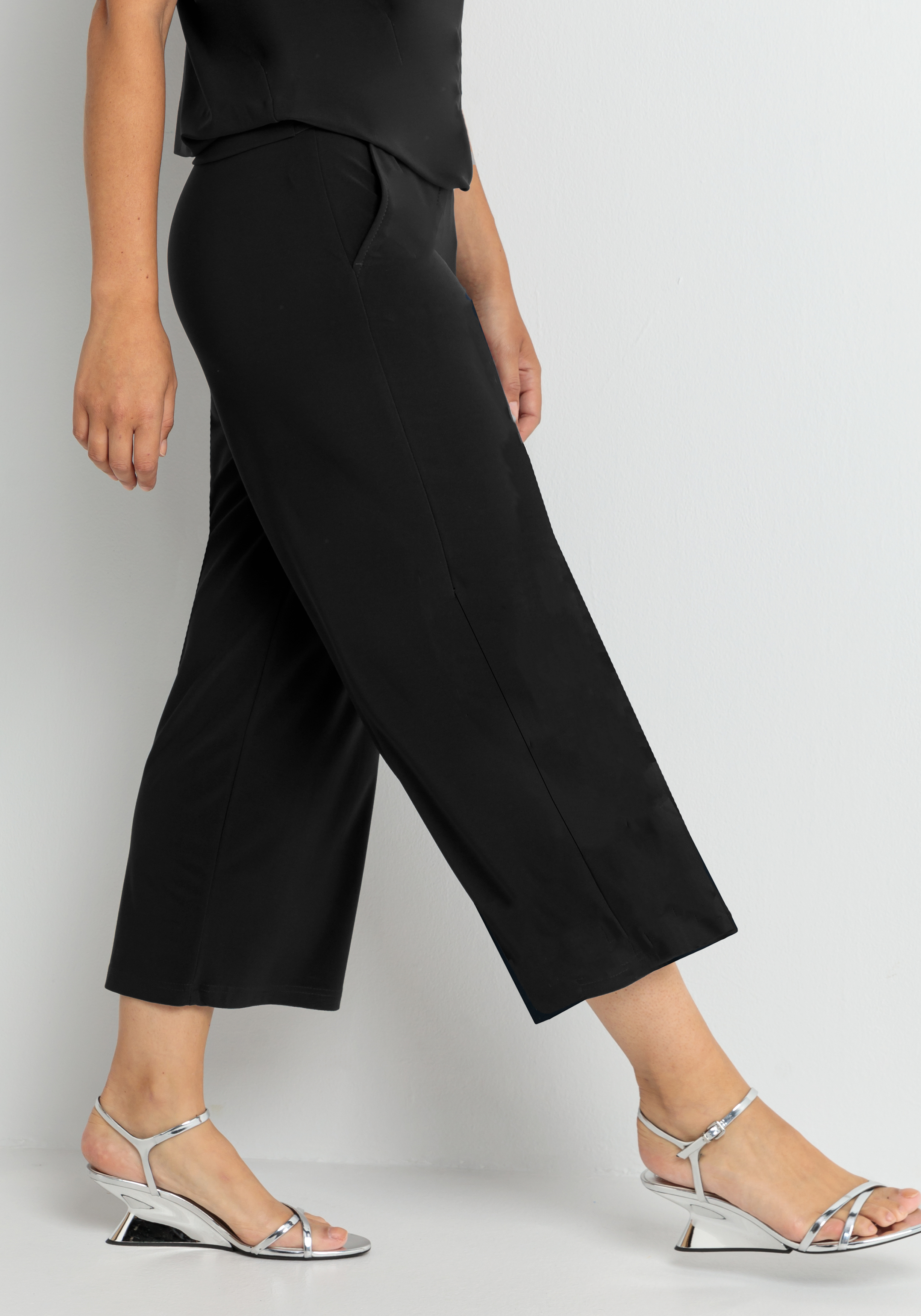 Eine Person, die eine schwarze, weit geschnittene Hose und silberne, offene Sandalen trägt, geht an einer schlichten, hellen Wand entlang. Nur die untere Hälfte ihres Körpers ist sichtbar.