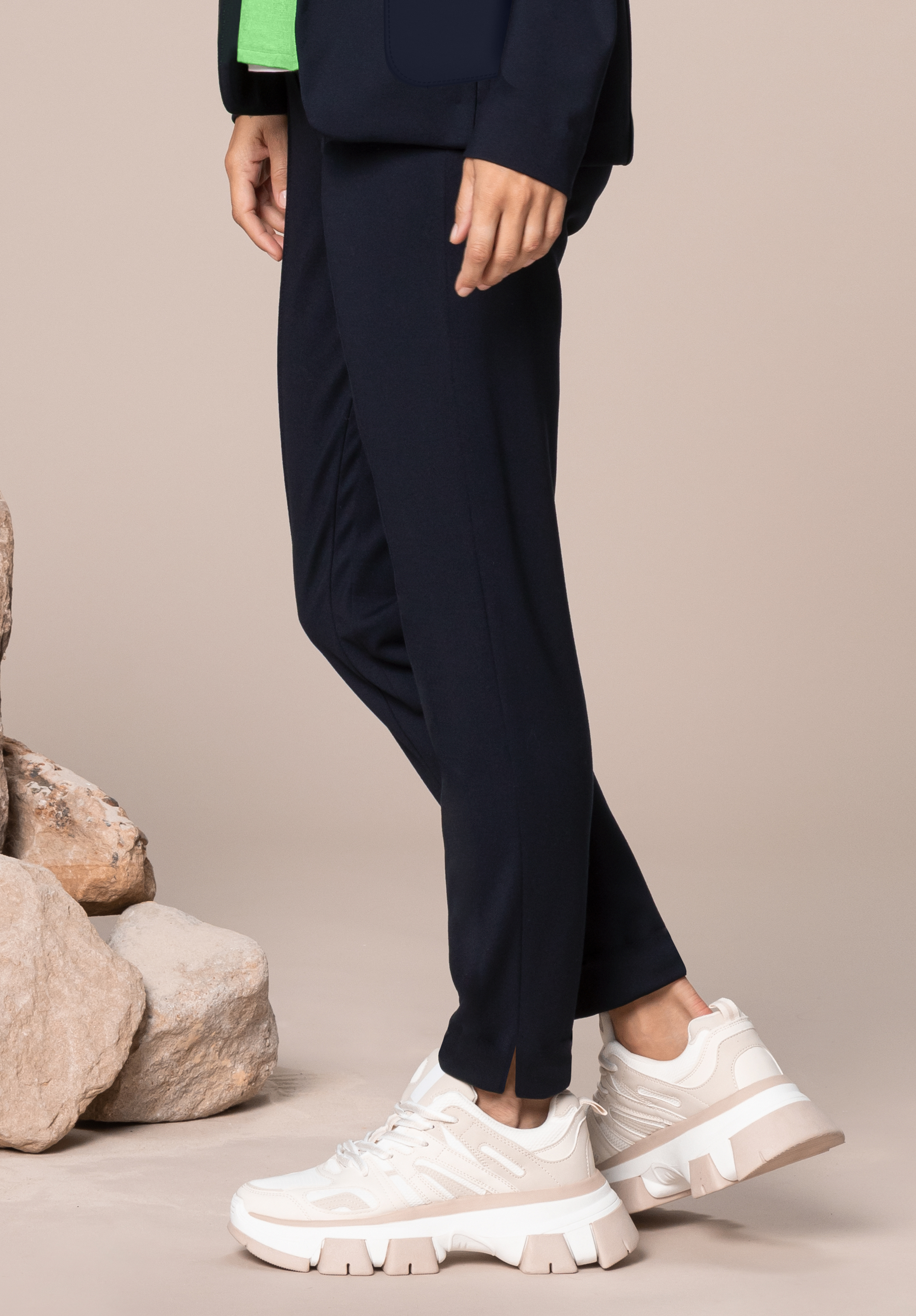 Eine Person in marineblauer Hose, dunkler Jacke und weißen klobigen Turnschuhen steht auf einem beigen Boden in der Nähe mehrerer großer, heller Felsen. Nur die untere Körperhälfte ist sichtbar.