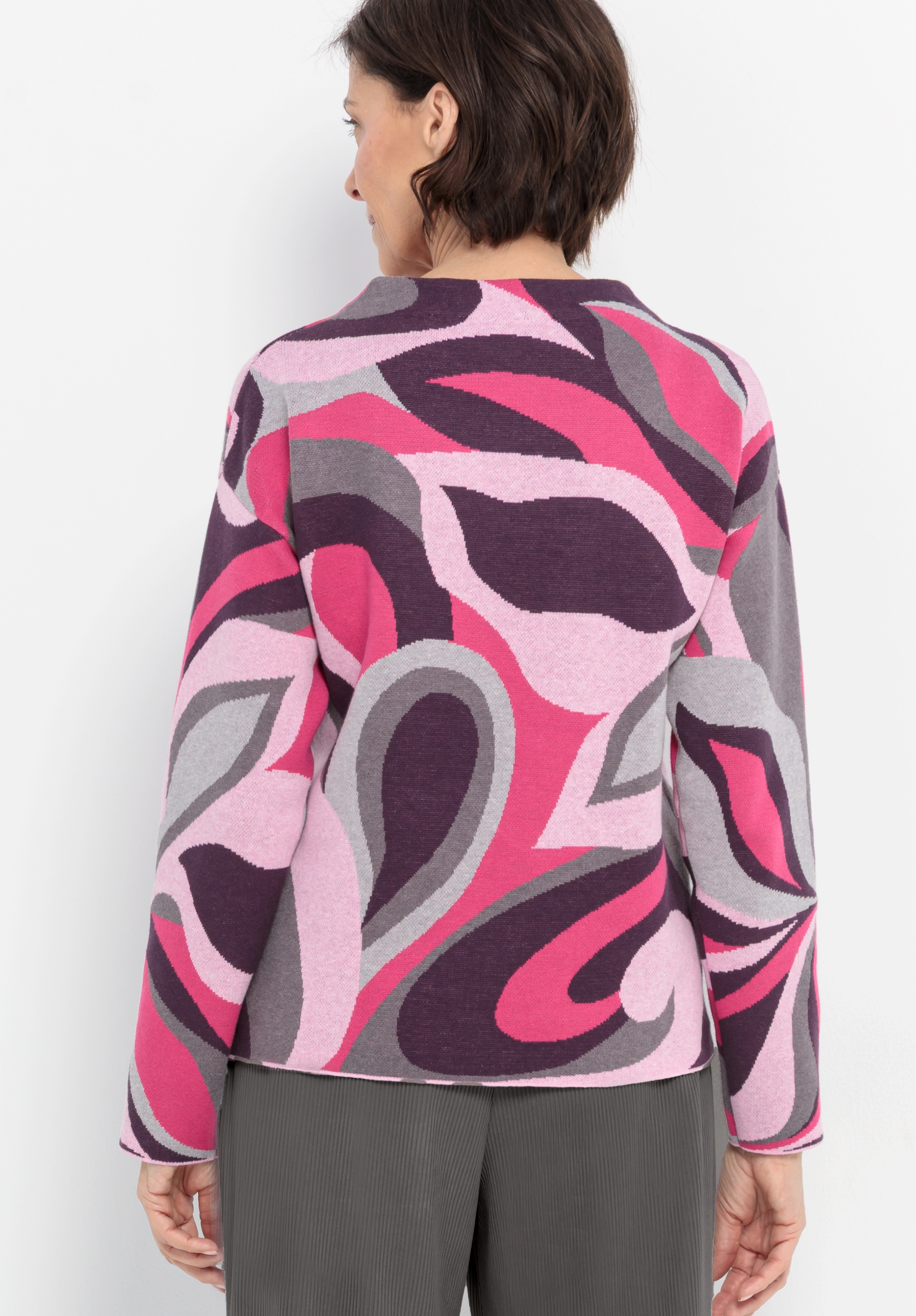 Eine Frau mit kurzen braunen Haaren ist von hinten zu sehen. Sie trägt einen langärmeligen Pullover mit einem auffälligen abstrakten Muster in Rosa-, Lila-, Grau- und Weißtönen und dazu eine graue Hose.