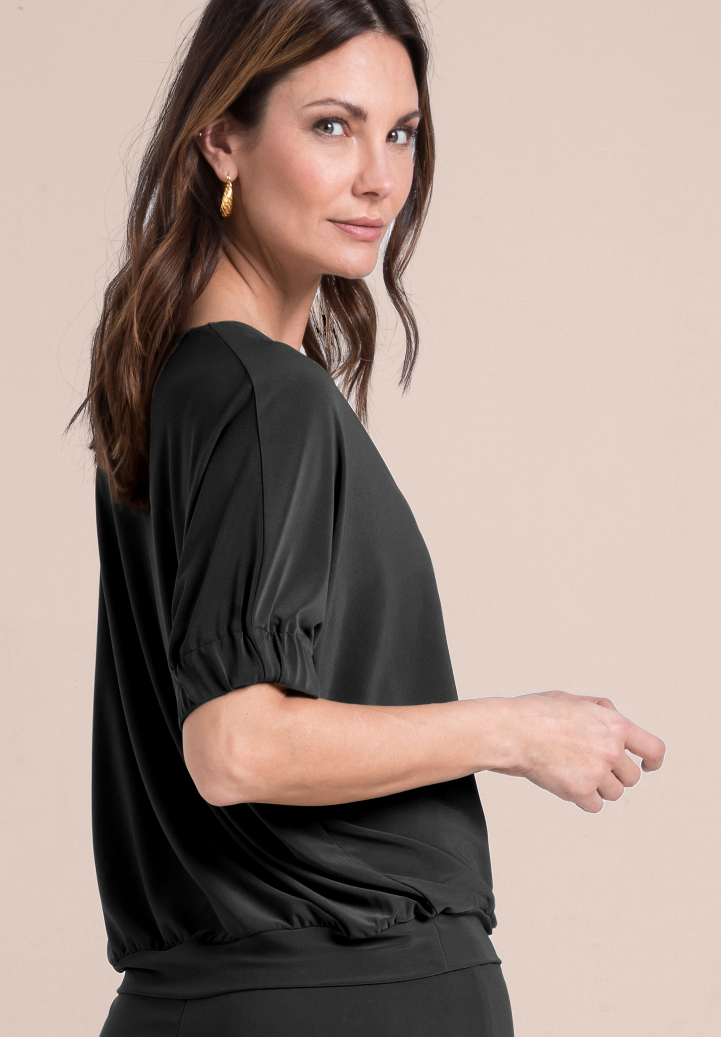 Eine Frau mit braunem Haar, die goldene Ohrringe und ein locker sitzendes schwarzes Oberteil mit gerafften Ärmeln trägt, steht seitlich vor einem einfarbig beigen Hintergrund und blickt mit einem leichten Lächeln in die Kamera.