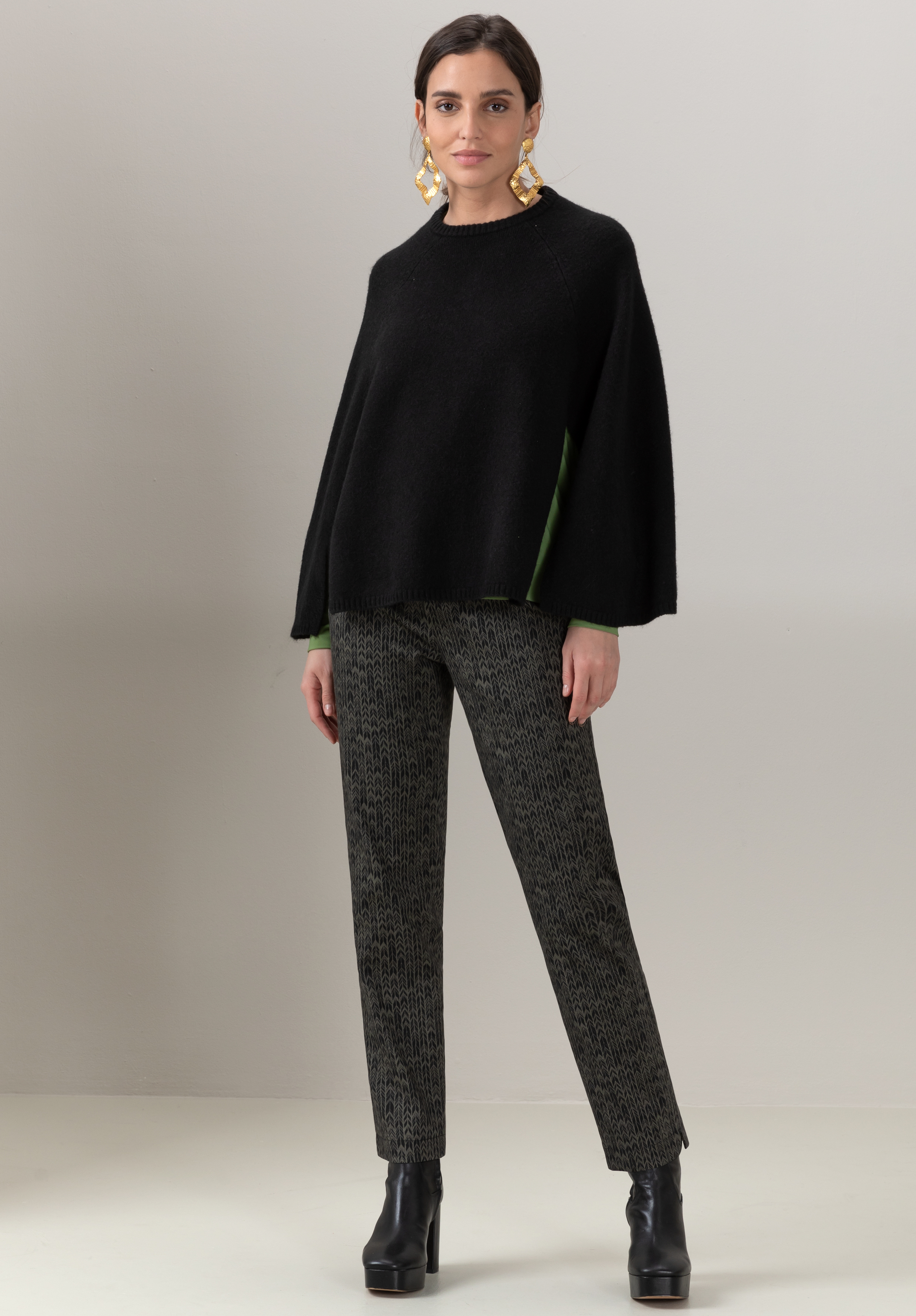 Eine Frau steht vor einer hellbeigen Wand. Sie trägt einen weiten schwarzen Pullover mit grünen Akzenten, gemusterte graue Hosen, schwarze Plateaustiefel und große Statement-Ohrringe.