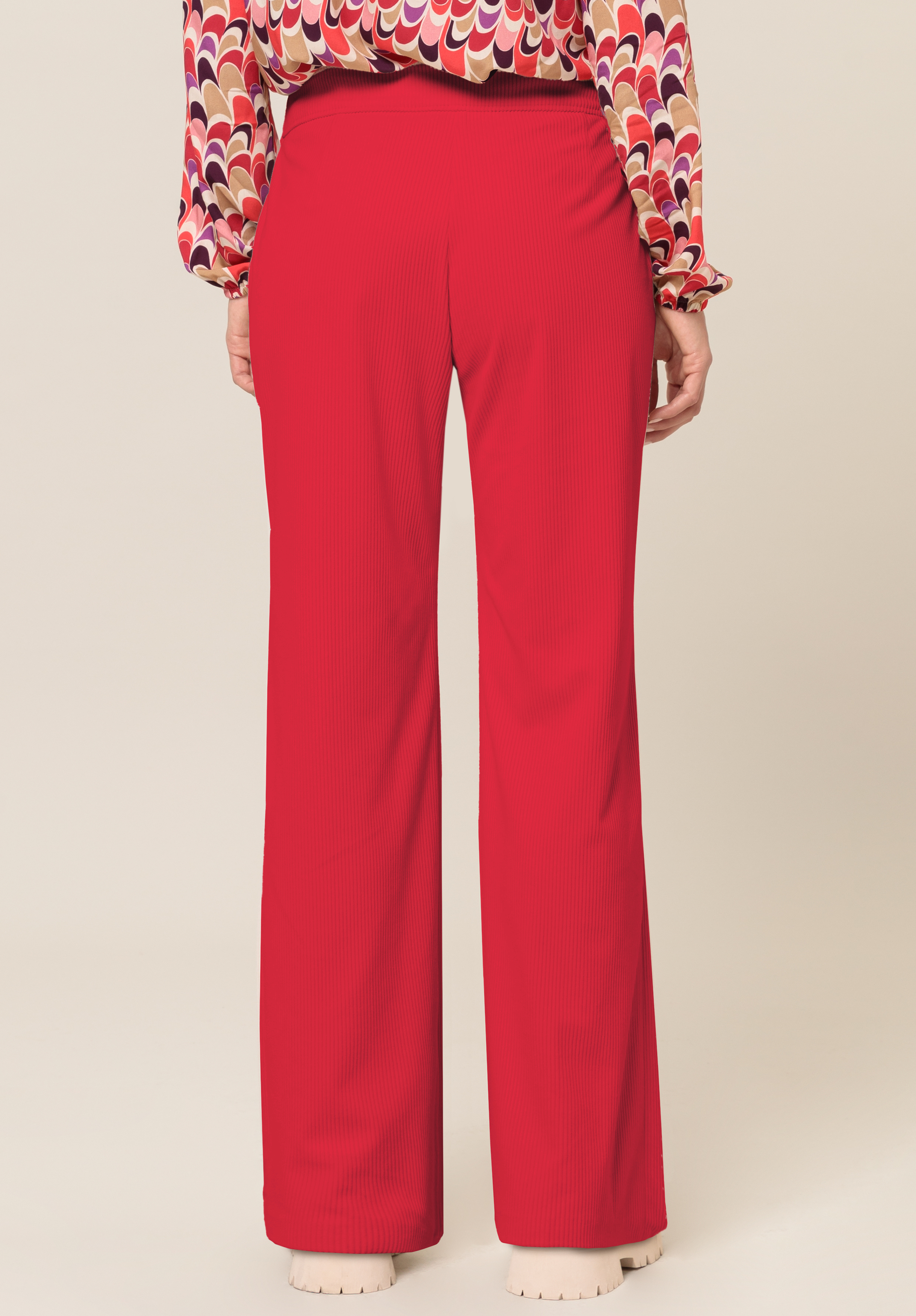 Eine Person trägt eine leuchtend rote Hose mit weitem Bein und eine bunt gemusterte Langarmbluse. Das Bild zeigt die Rückansicht von der Taille bis zu den Füßen, wobei cremefarbene Schuhe sichtbar sind.