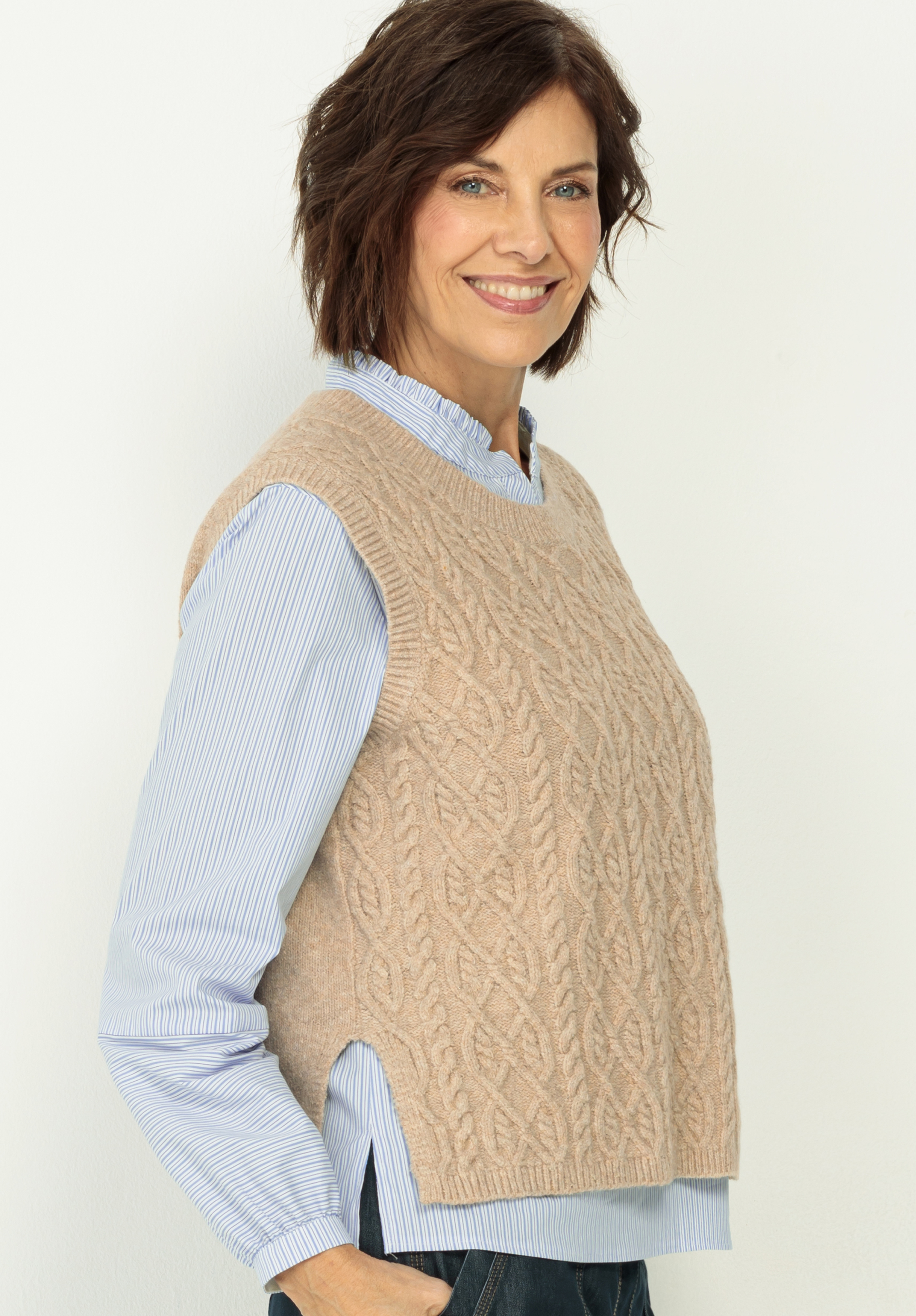 Eine Frau mit kurzen braunen Haaren lächelt, während sie ein hellblau gestreiftes Hemd unter einer beigen Strickweste trägt und vor einem schlichten hellen Hintergrund steht.