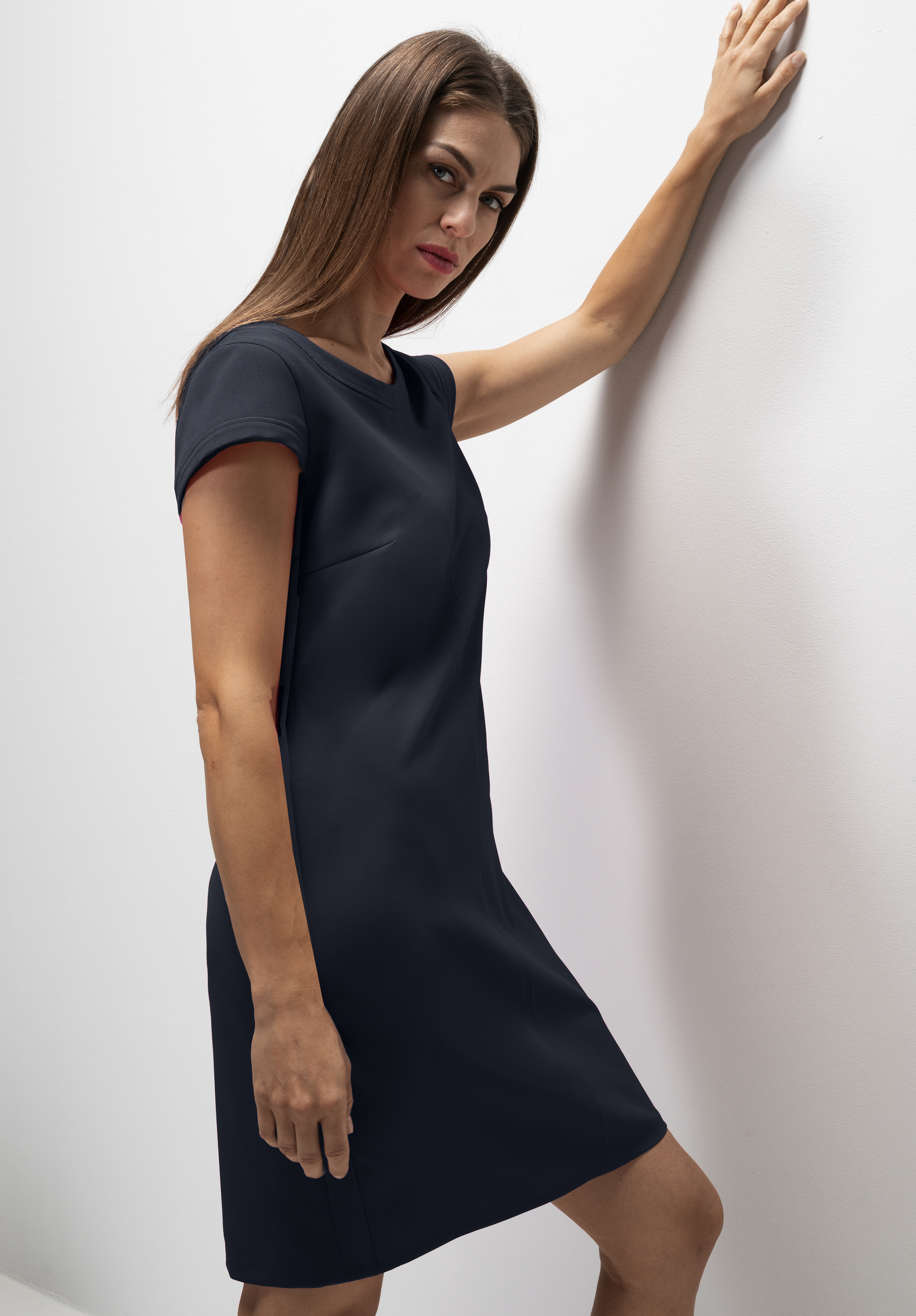 Eine Frau mit glattem braunem Haar trägt ein kurzärmeliges, knielanges schwarzes Kleid. Sie steht vor einer weißen Wand, stützt sich mit einem Arm auf der Wand ab und blickt mit neutralem Blick in die Kamera.